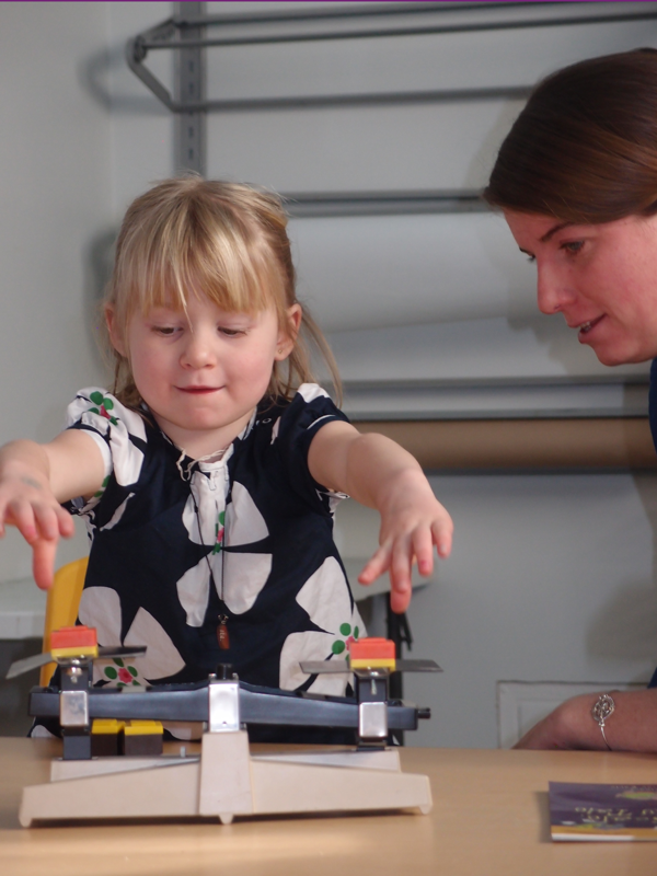 A young girl with blonde hair reaches out to interact with abalance scale on a table, while a woman looks on nearby.