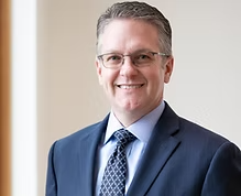A professional man in a dark suit, light blue shirt, and patterned tie, smiling in an office setting with beige walls and a window in the background. Doug Langen Director on the board of directors