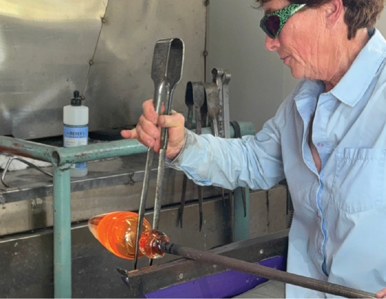 A woman wearing safety glasses and a light blue shirt using tongs to shape molten glass in a glassblowing studio.