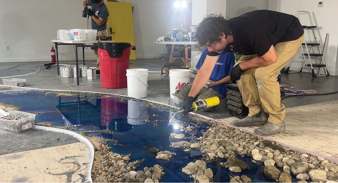 A person working on a small indoor artificial river installation, using a torch to create a textured water effect, with construction supplies and another person in the background.