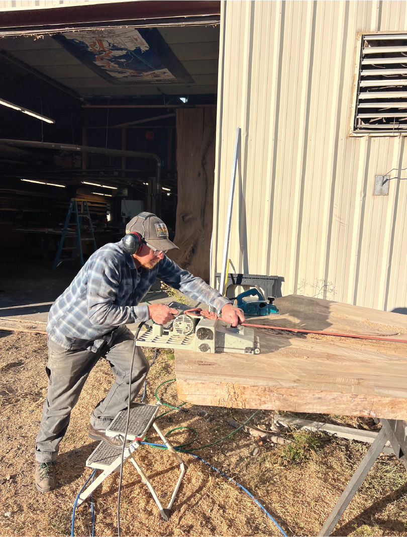 A man wearing a baseball cap, safety glasses, and ear protection uses a power planer on a large piece of wood outside a building. The woodworking setup includes a portable workbench with tools and cords, with sunlight illuminating the scene.