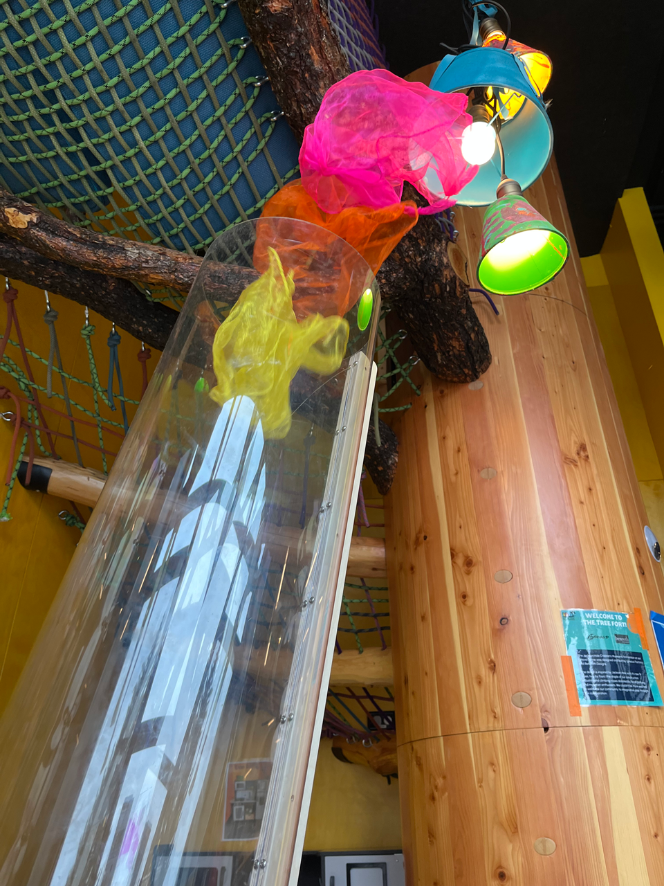 Indoor play area with colorful hanging lamps, a tree branch, and rope climbing structures, featuring wooden surfaces and a netted climbing area.