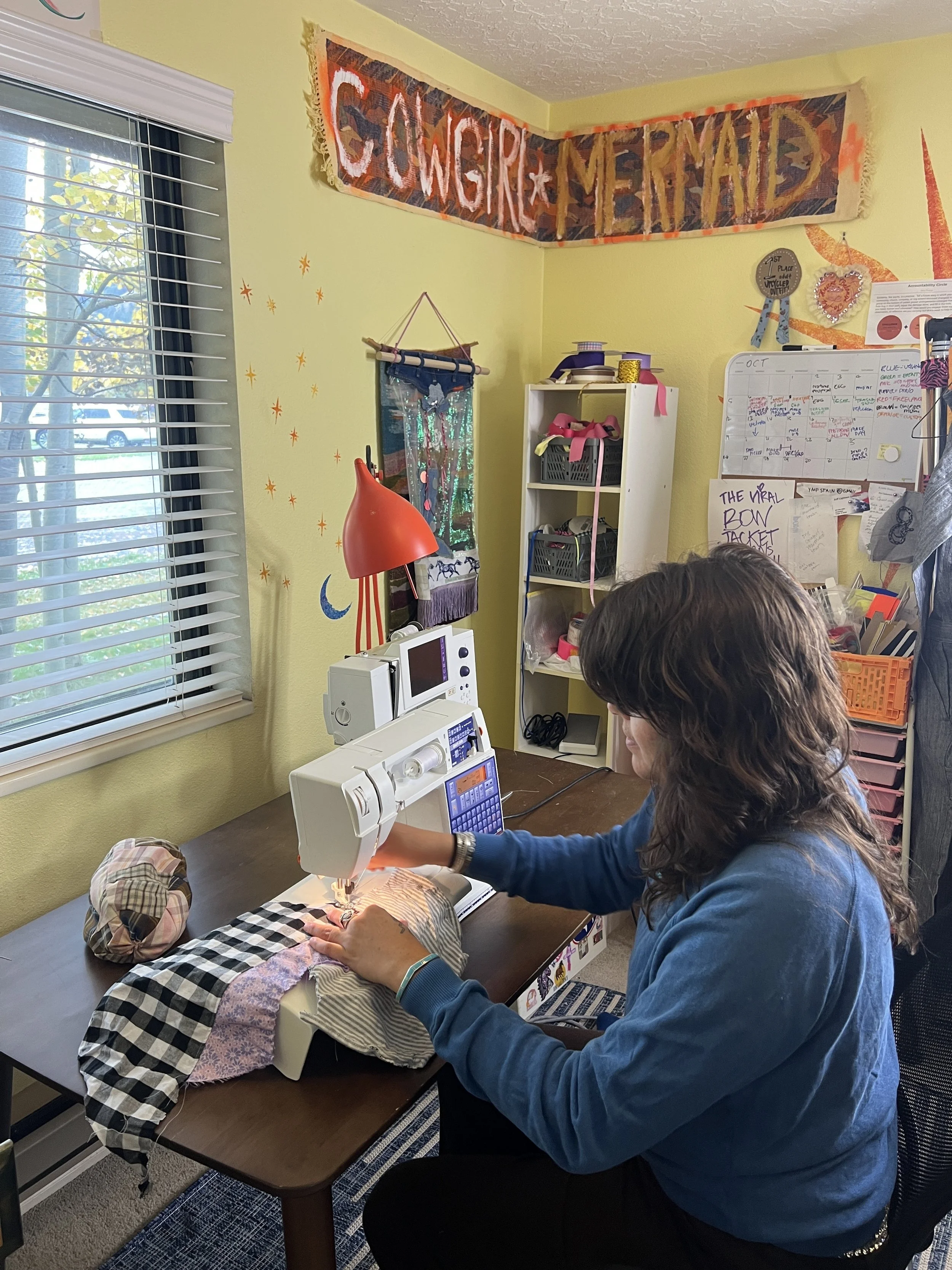 Woman sewing fabric pieces on a white sewing machine in a cozy home craft room with yellow walls, decorated with a 'CONGRATS MERMAID' banner, star and moon wall decals, and colorful storage bins.