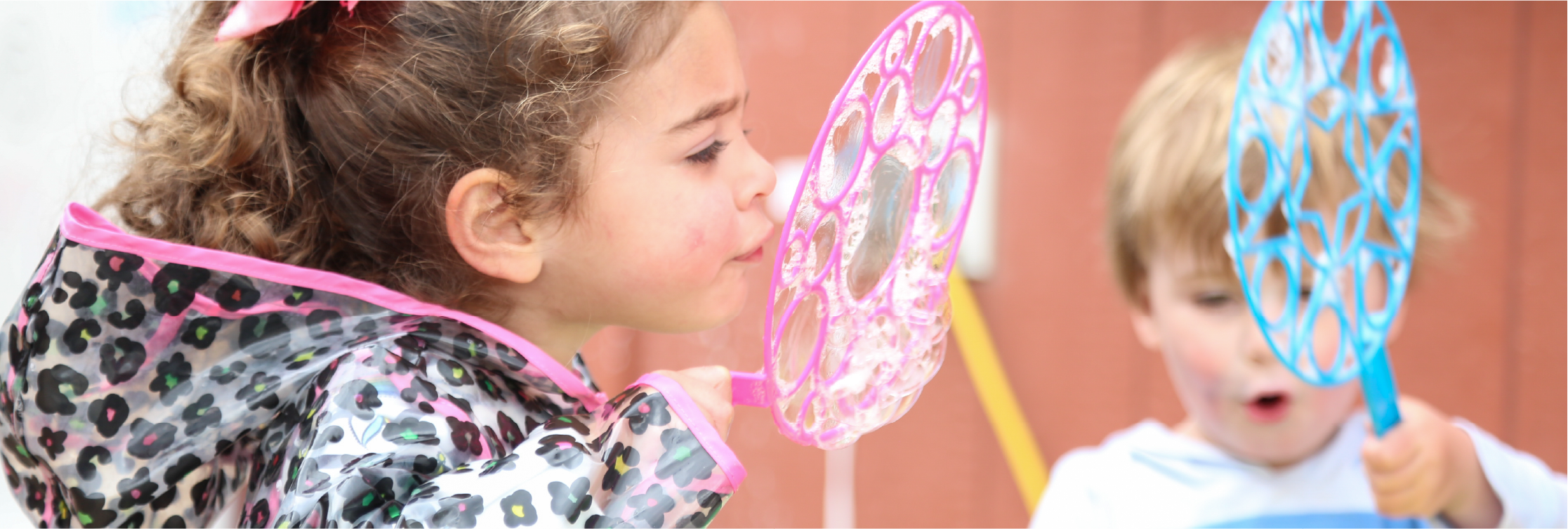 Two children playing with bubble wands indoors. The girl in the foreground has curly hair, is wearing a leopard print jacket with pink trim, and is blowing bubbles. The boy in the background has straight hair, is holding a blue bubble wand, and is watching the bubbles.