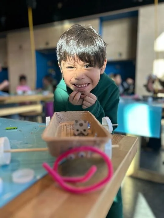 Child smiling happily at a table with a small device featuring white dots, in a classroom or play area with other children in the background.