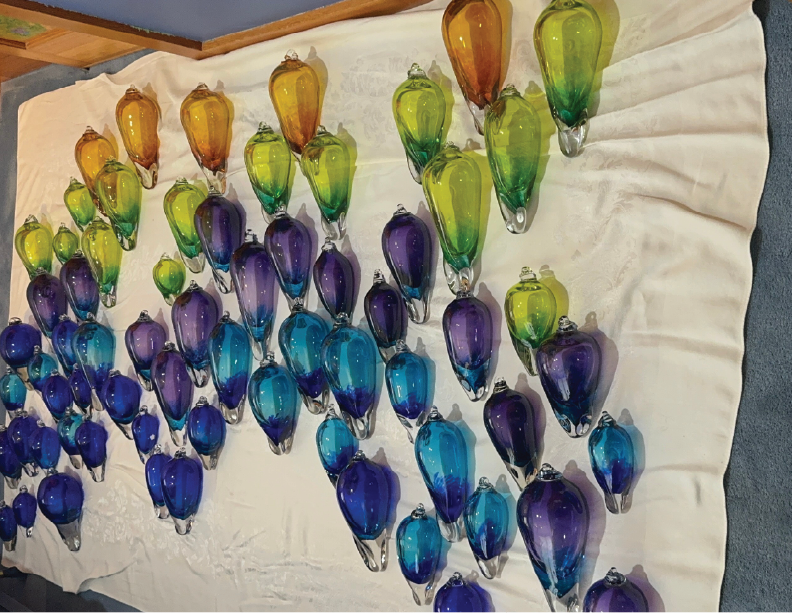 A large collection of glass art ornaments in shades of yellow, green, blue, purple, and orange arranged on a white cloth surface.