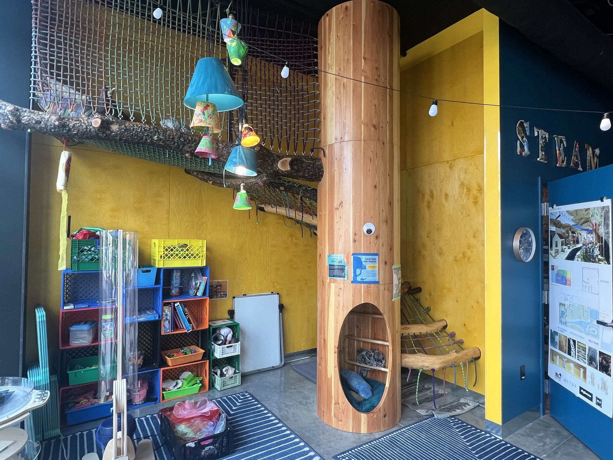 Indoor play area with yellow and blue walls, a wooden climbing structure with a circular opening, colorful hanging lights, storage shelves with toys and books, and a mural or sign reading 'STEAM' on the wall.