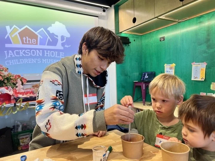 A young adult demonstrating a science experiment with two young children at a table in a museum classroom, with a colorful logo and green wall in the background.