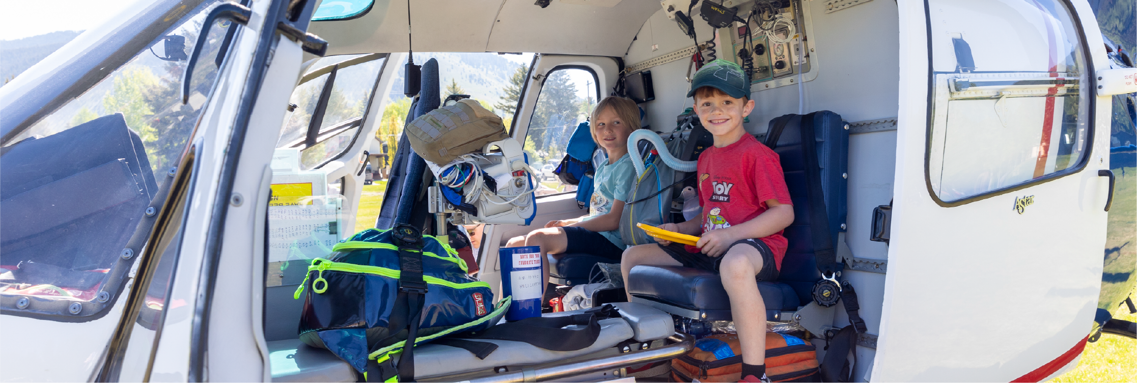 Two children sitting inside a helicopter, surrounded by various equipment and backpacks, during daytime outdoors.