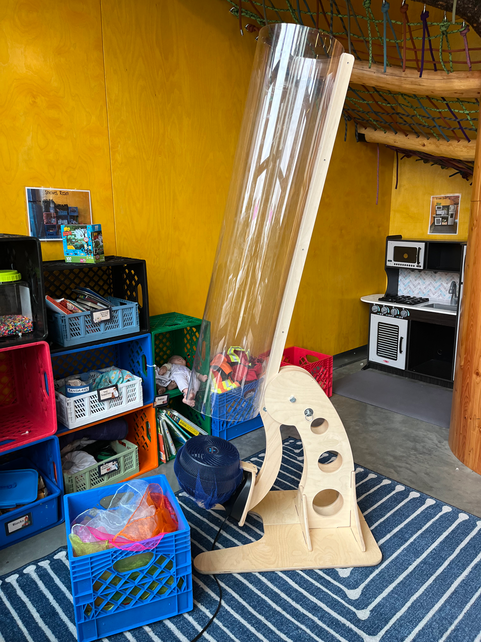 Indoor children’s play area with colorful storage bins filled with toys, a wooden climbing structure with a slide, a play kitchen set, and a blue striped rug on the floor.