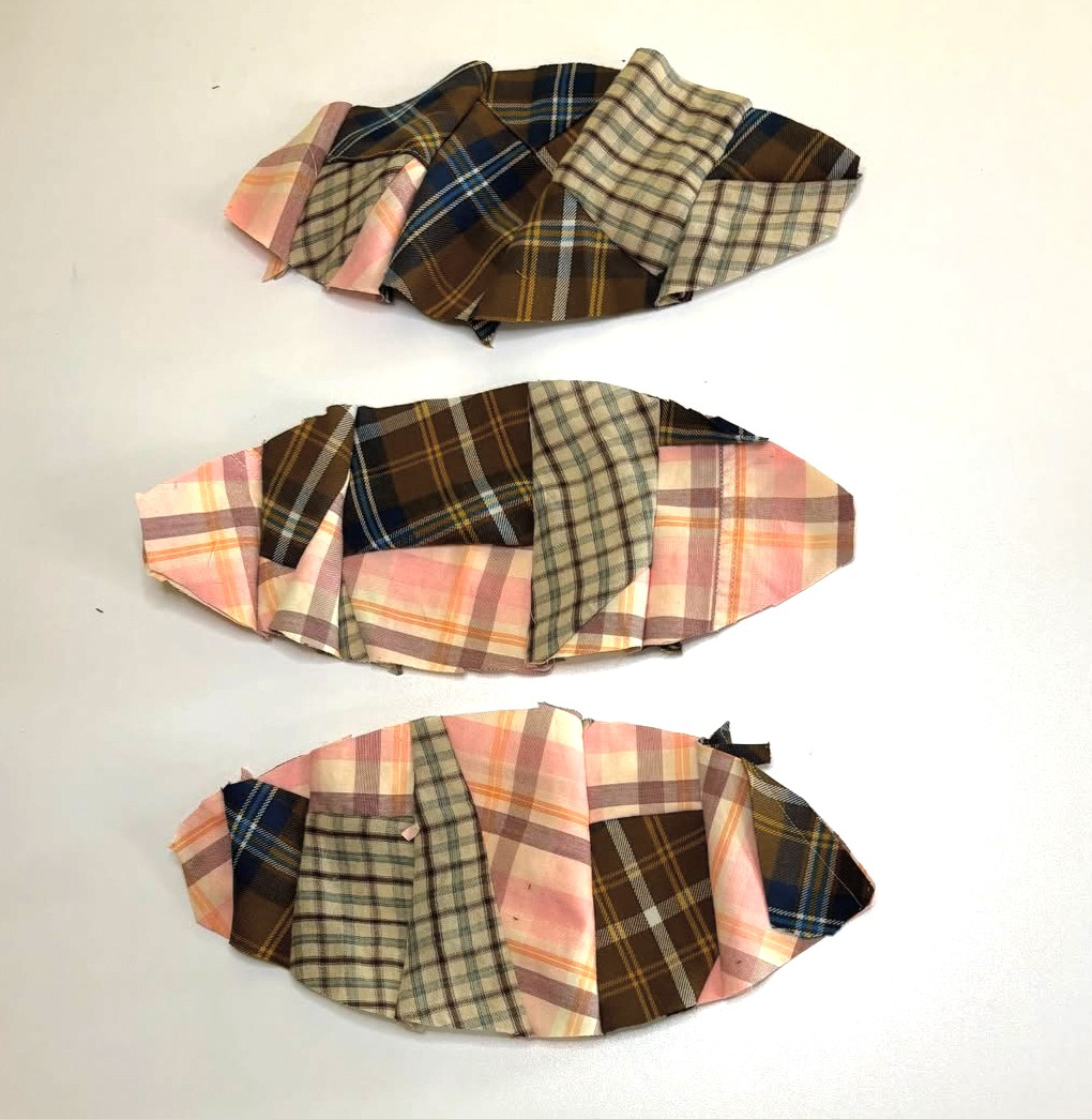 Three pieces of fabric fashioned into fish shapes, made from patchwork plaid cloths in various colors including pink, brown, and beige, arranged on a white surface.