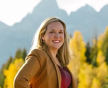 Smiling woman with blonde hair standing outdoors near yellow trees and mountains.