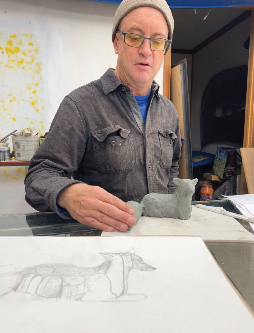 A man wearing glasses, a gray beanie, and a gray shirt, working on a small gray clay sculpture of an alpaca at a desk. Sketches are visible on the paper in front of him.