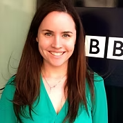 A young woman with long brown hair, smiling, wearing a teal top, standing in front of a BBC sign. Grace Johnston Director on the board of directors
