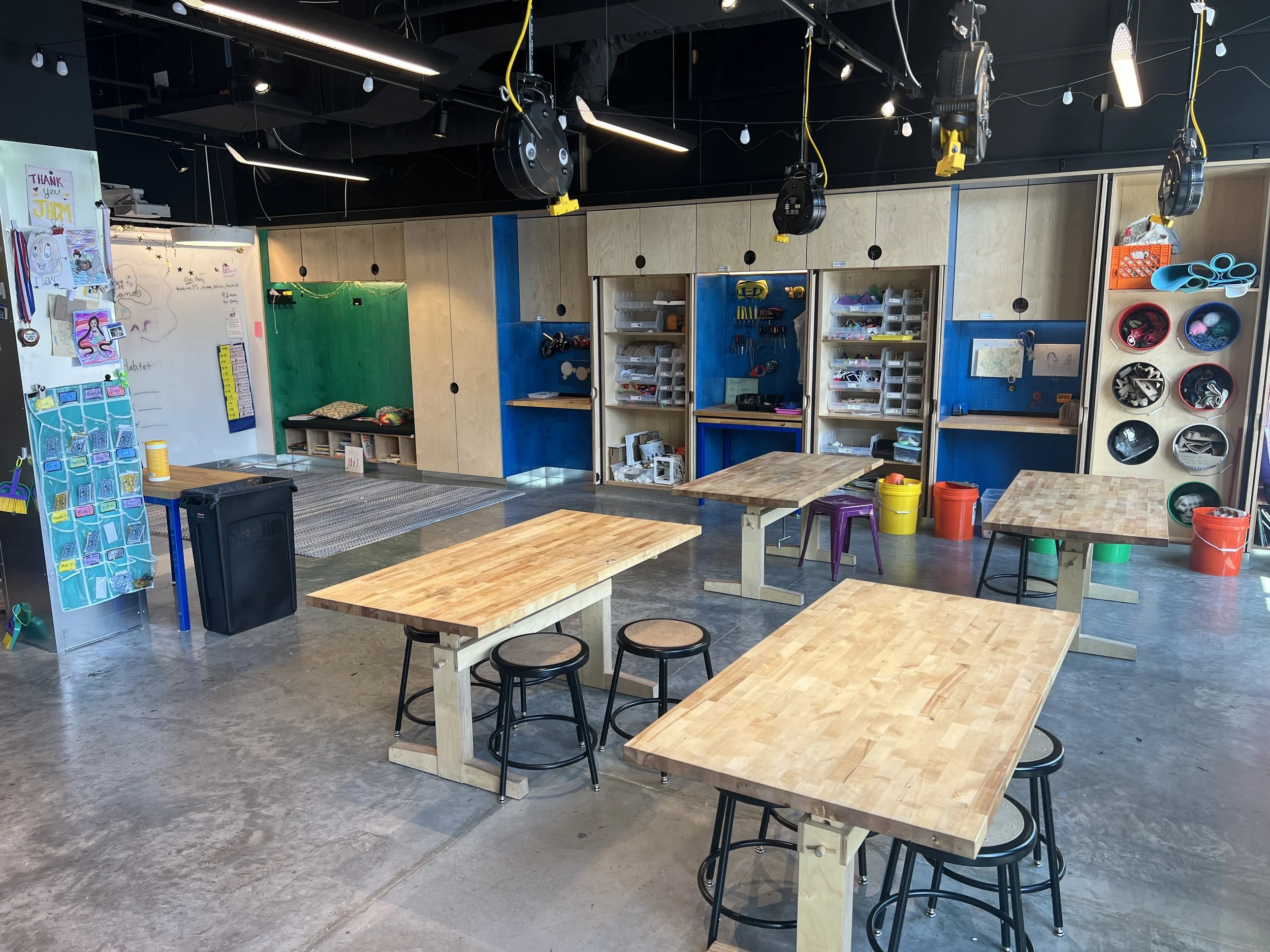 An art classroom with wooden tables and black stools, colorful storage bins and shelves filled with supplies, a whiteboard with drawings, and a small green and blue section with hanging tools.