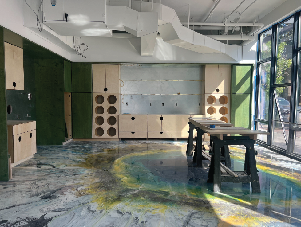 Interior space under construction with green and beige cabinetry, a colorful swirling floor, and large floor-to-ceiling windows on the right, showing outdoor trees.