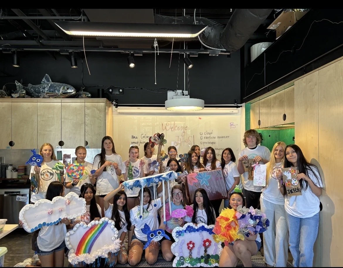 A group of young girls in a room holding colorful, craft-made clouds, rainbows, and other decorative items, in front of a whiteboard with writing and a large fish sculpture on top of the cabinets.