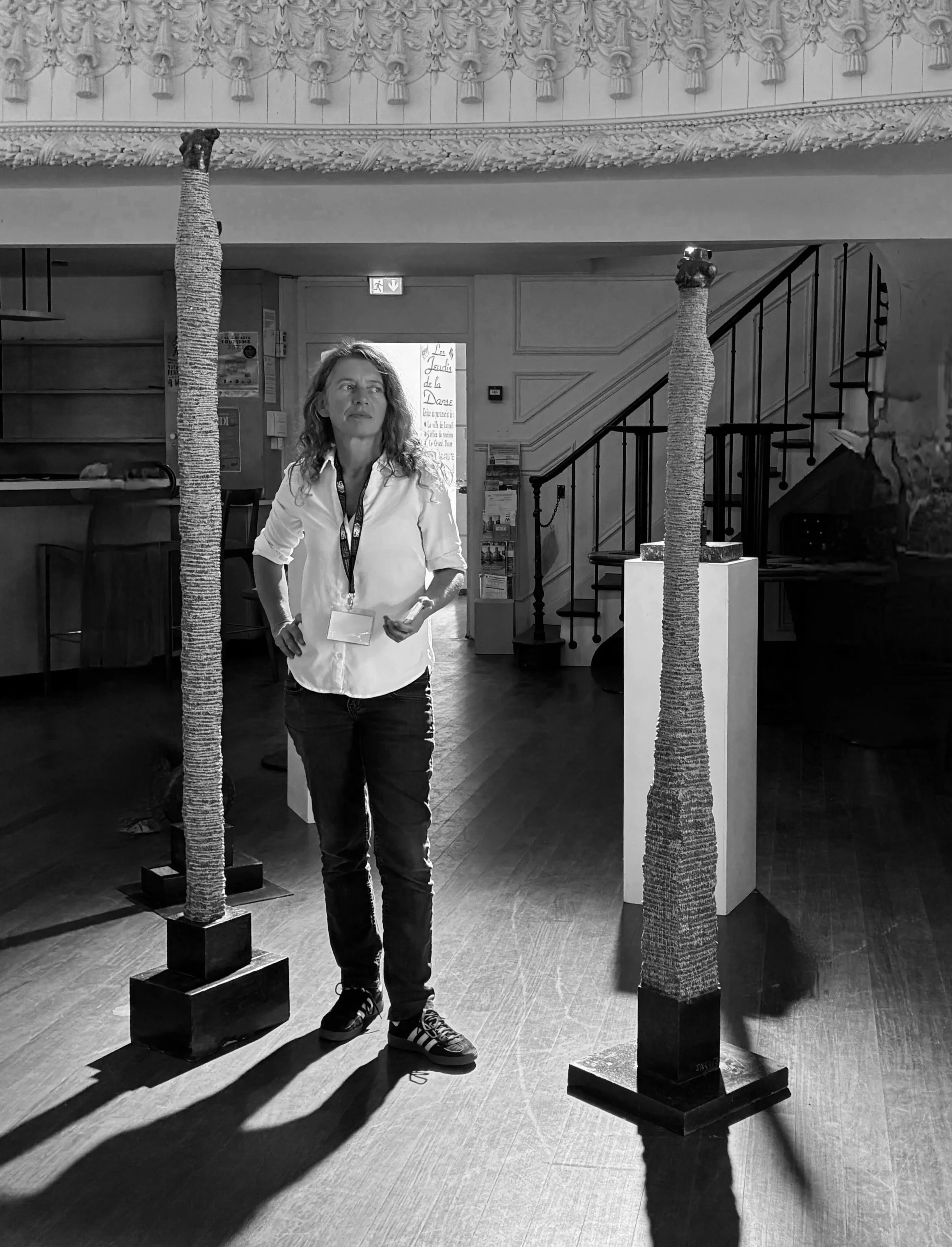 Artiste de sculpture contemporaine dans une galerie d’art, entourée de deux sculptures verticales en pierre. Œuvres longilignes, installation contemporaine, exposition d’art, sculpture sur pierre, créations originales en granite, marbre ou calcaire.