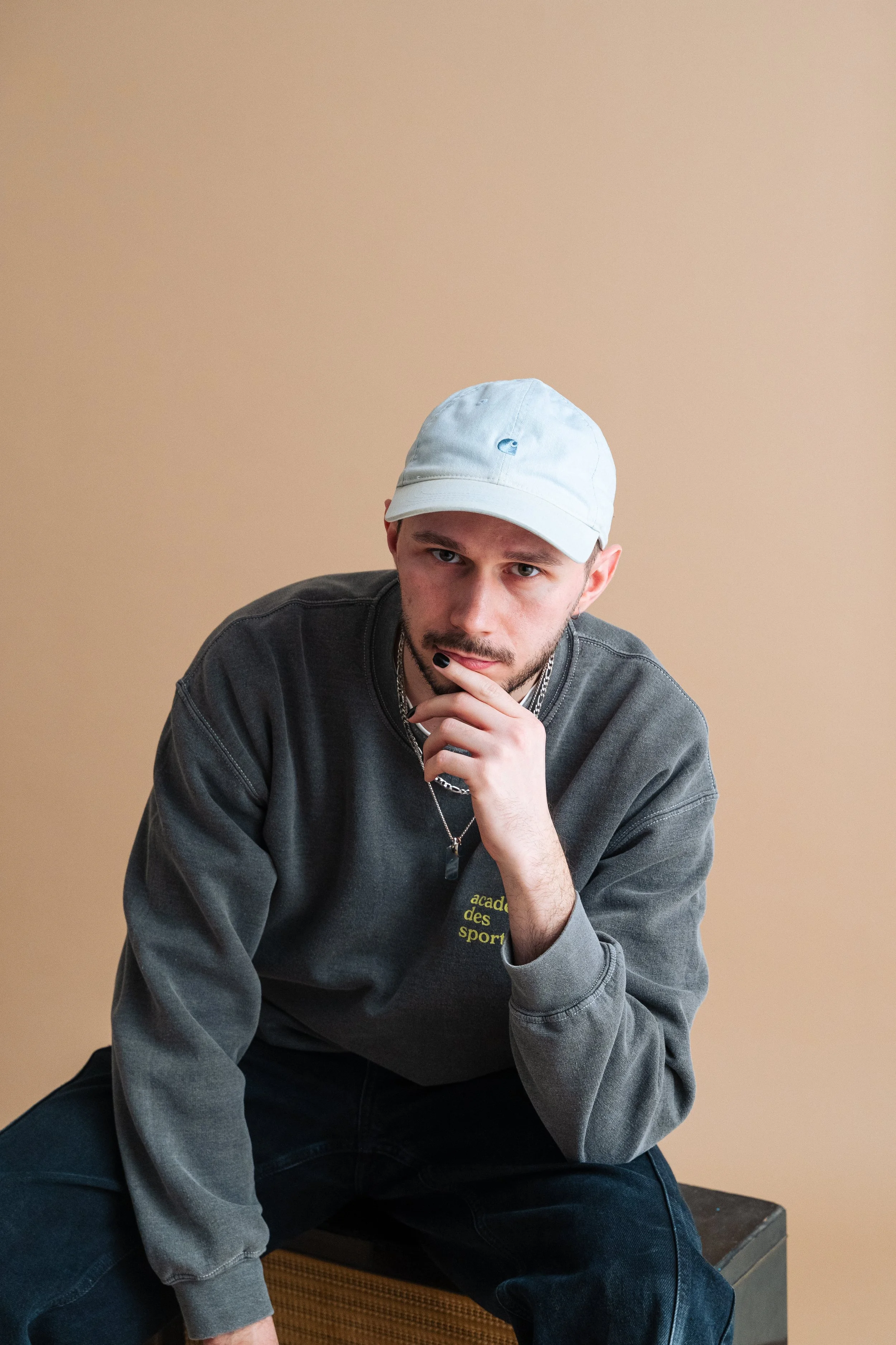 Ein junger Mann trägt eine weiße Kappe und ein graues Sweatshirt, sitzt auf einem Hocker vor einem beigen Hintergrund, mit nachdenklicher Miene und der Hand am Kinn.
