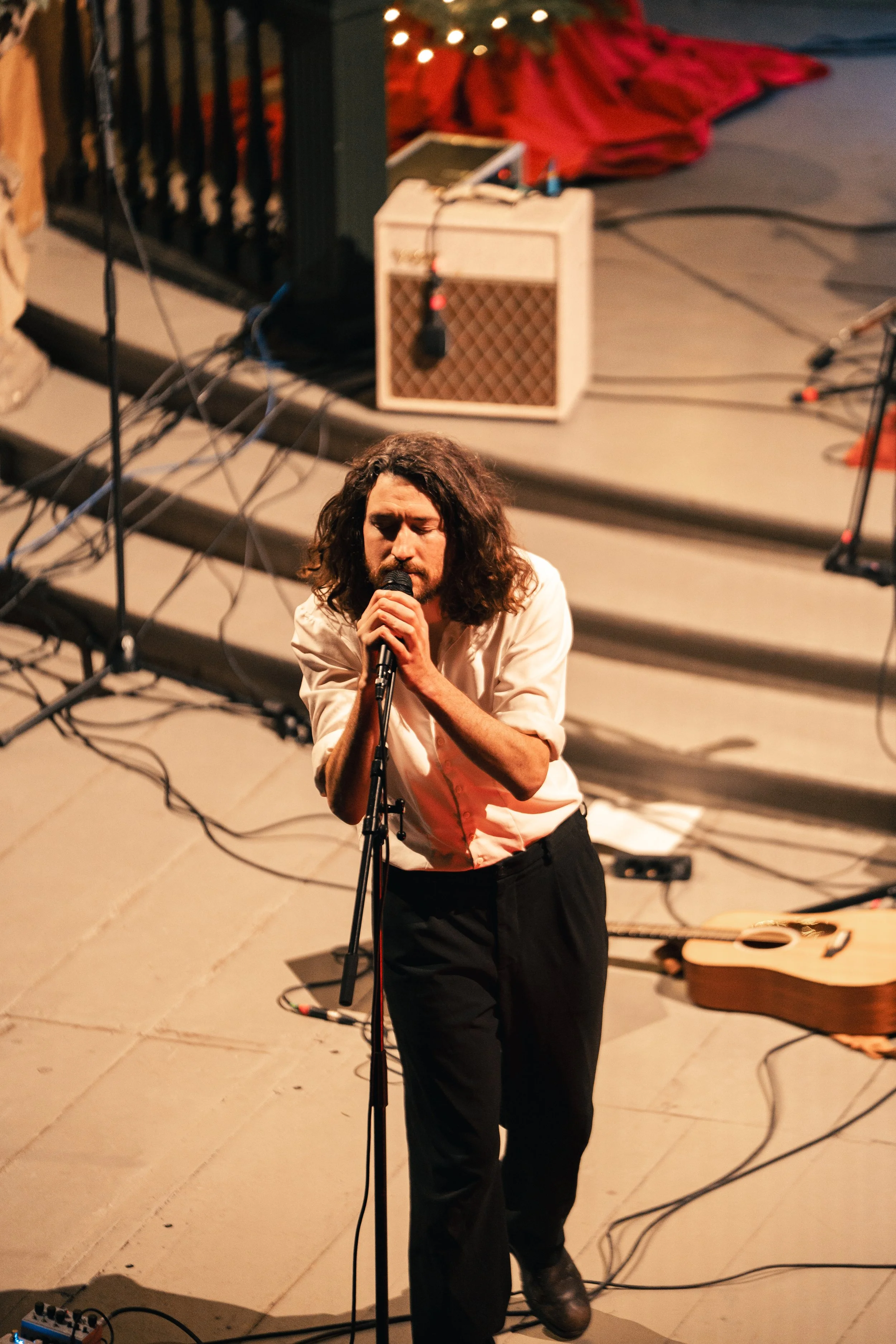 Ein Mann mit langem, lockigem Haar singt mit ruhigem Ausdruck in einem Mikrofon auf einer Bühne, umgeben von Musikanlage und Kabeln.