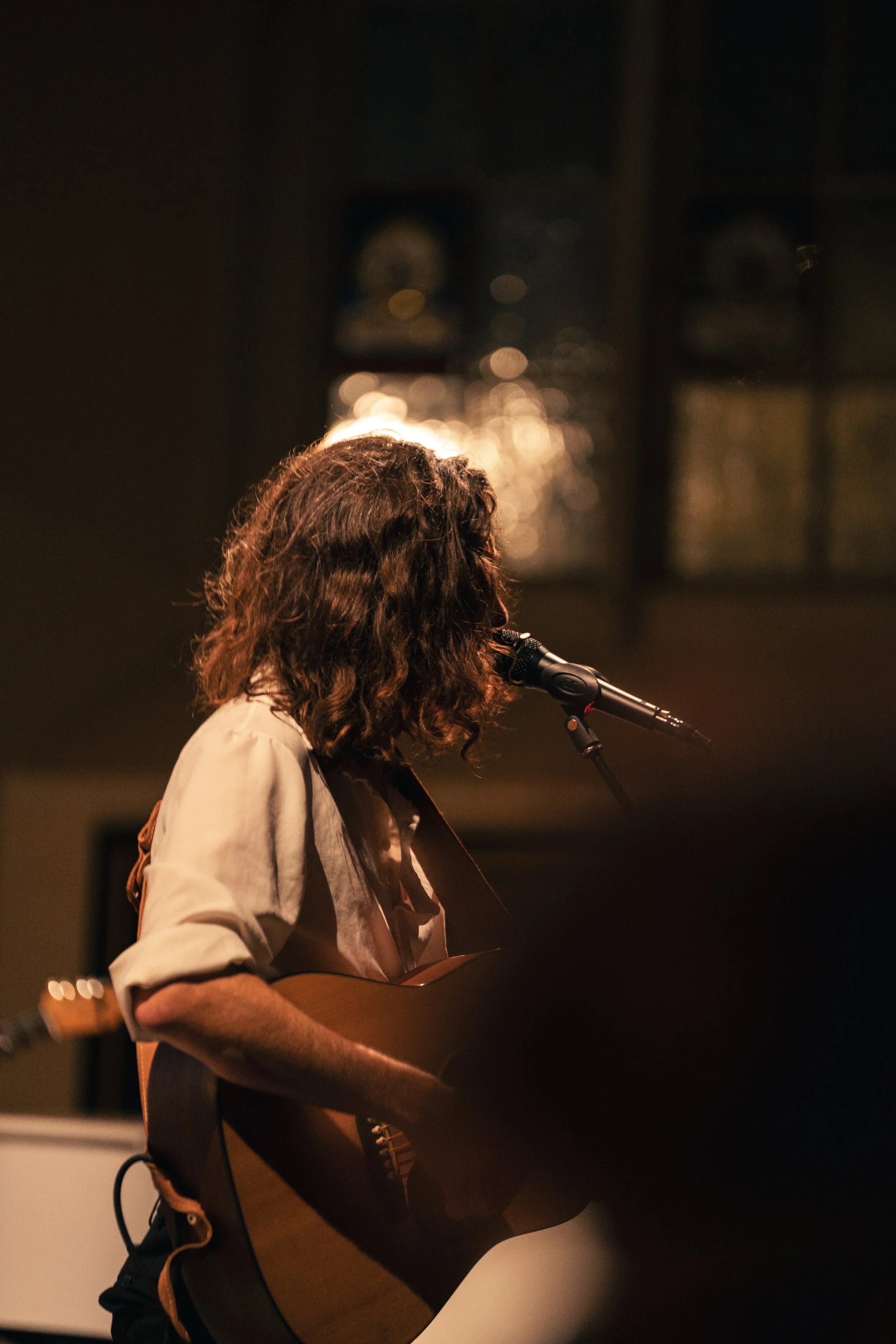 Frau mit wilden braunen Haaren spielt Gitarre und singt an einem Mikrofon in einem dunklen Raum mit warmem Licht.