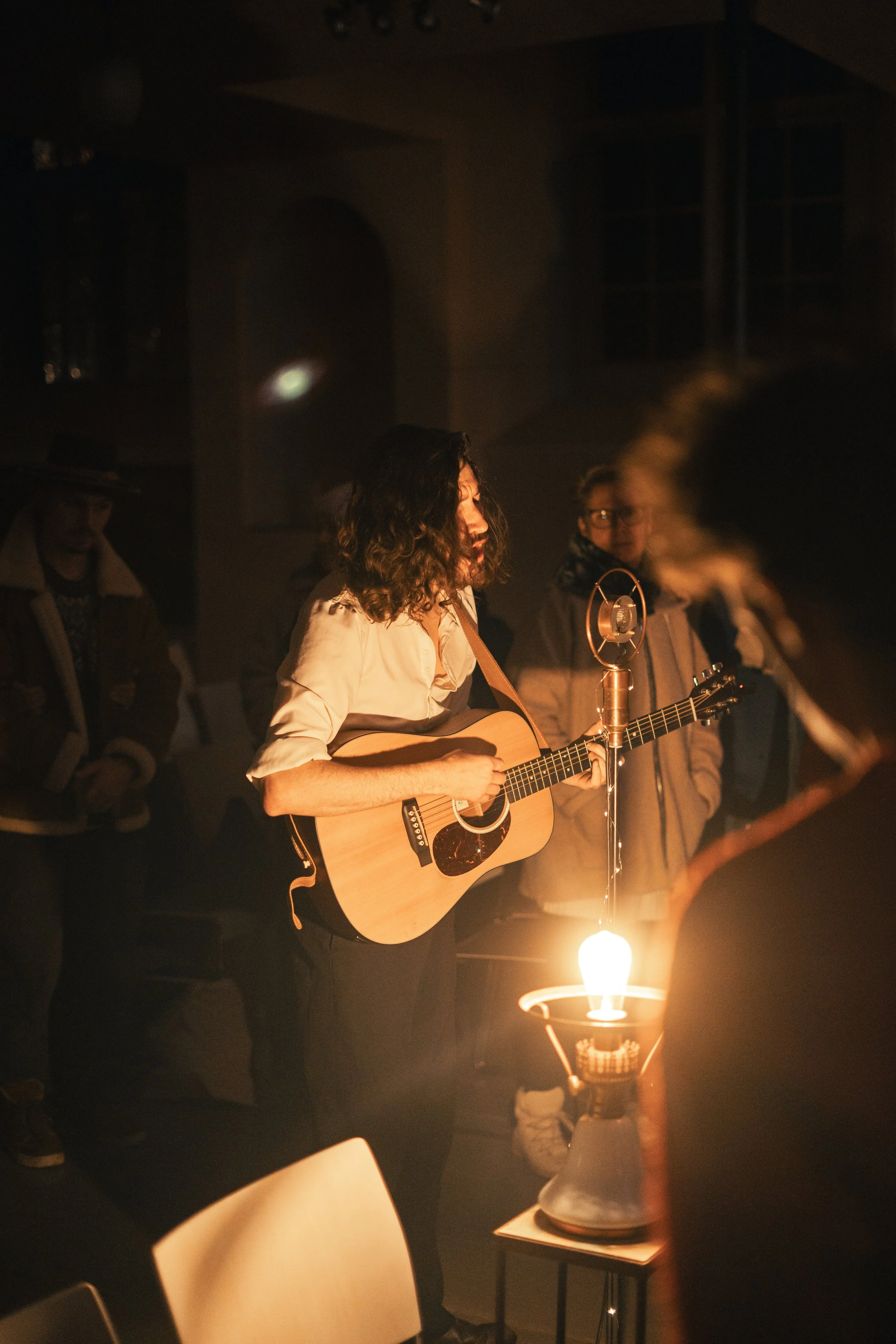 Eine Person spielt Gitarre in einem dunklen Raum, beleuchtet von einer Vintagelampe, mit mehreren im Hintergrund stehenden Menschen.