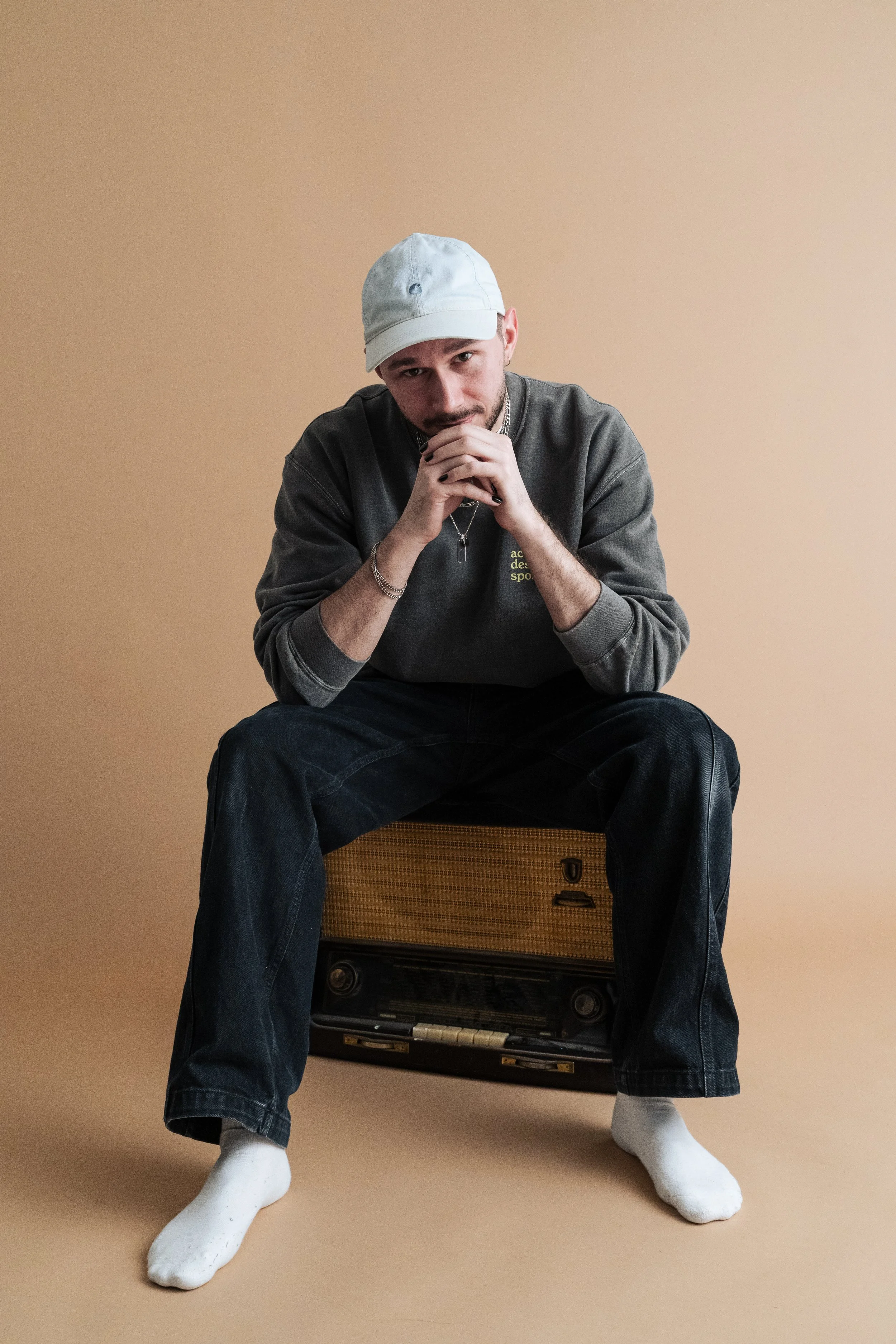 Junger Mann sitzt auf einem alten Radio gegen einen beige Hintergrund, trägt eine weiße Mütze, grauen Pullover, schwarze Jeans, weiße Socken und schaut nach vorne.