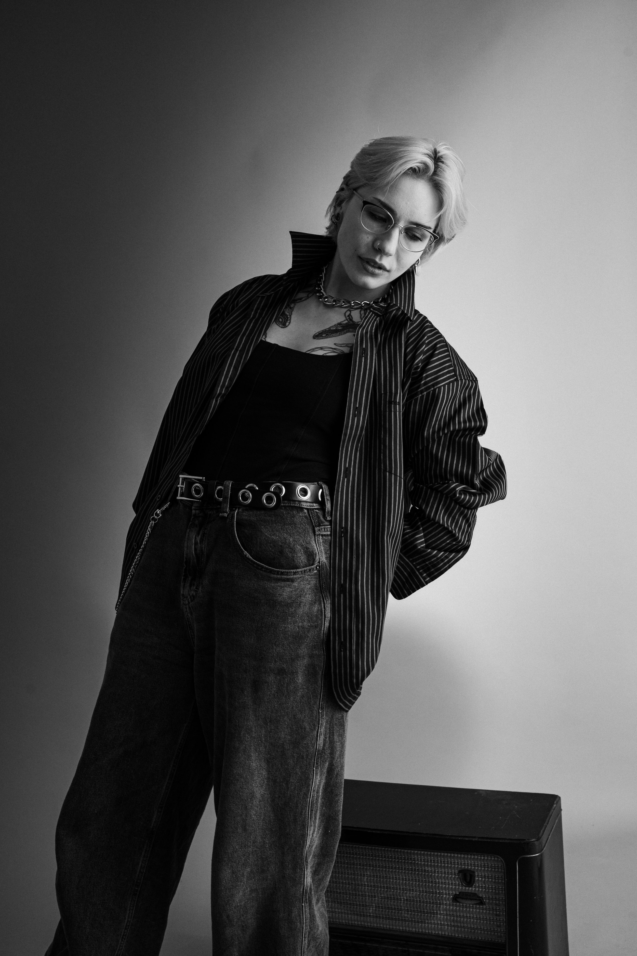 Schwarz-Weiß-Foto einer jungen Frau mit kurzen, blonden Haaren, die eine dunkle, offene Jacke trägt. Sie steht vor einem einfachen Hintergrund mit einem kleinen Verstärker oder Lautsprecher auf dem Boden neben ihr.