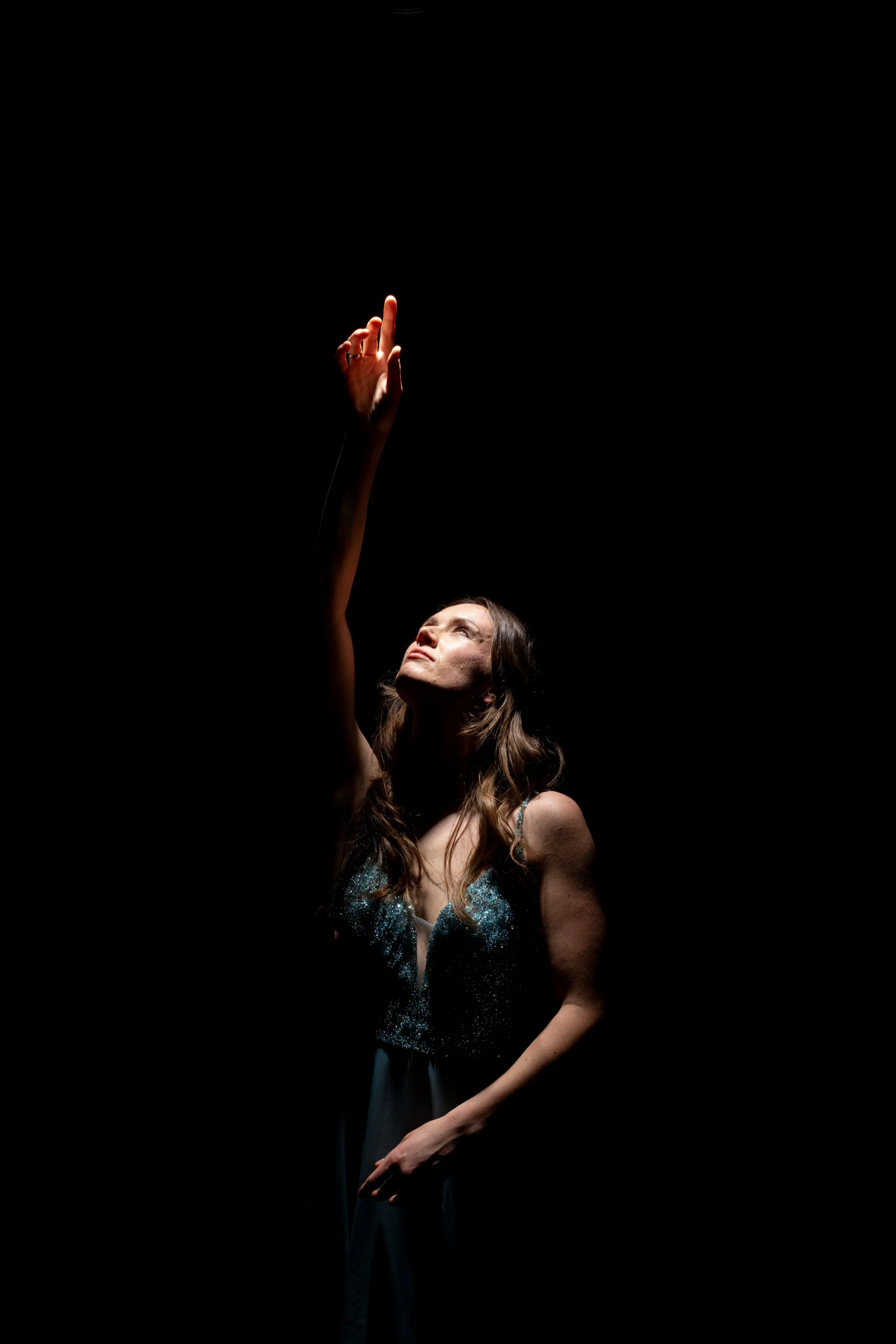 Eine Frau in einem eleganten Kleid hebt die Hand zum Himmel, beleuchtet durch einen fokussierten Lichtstrahl, vor einem schwarzen Hintergrund.