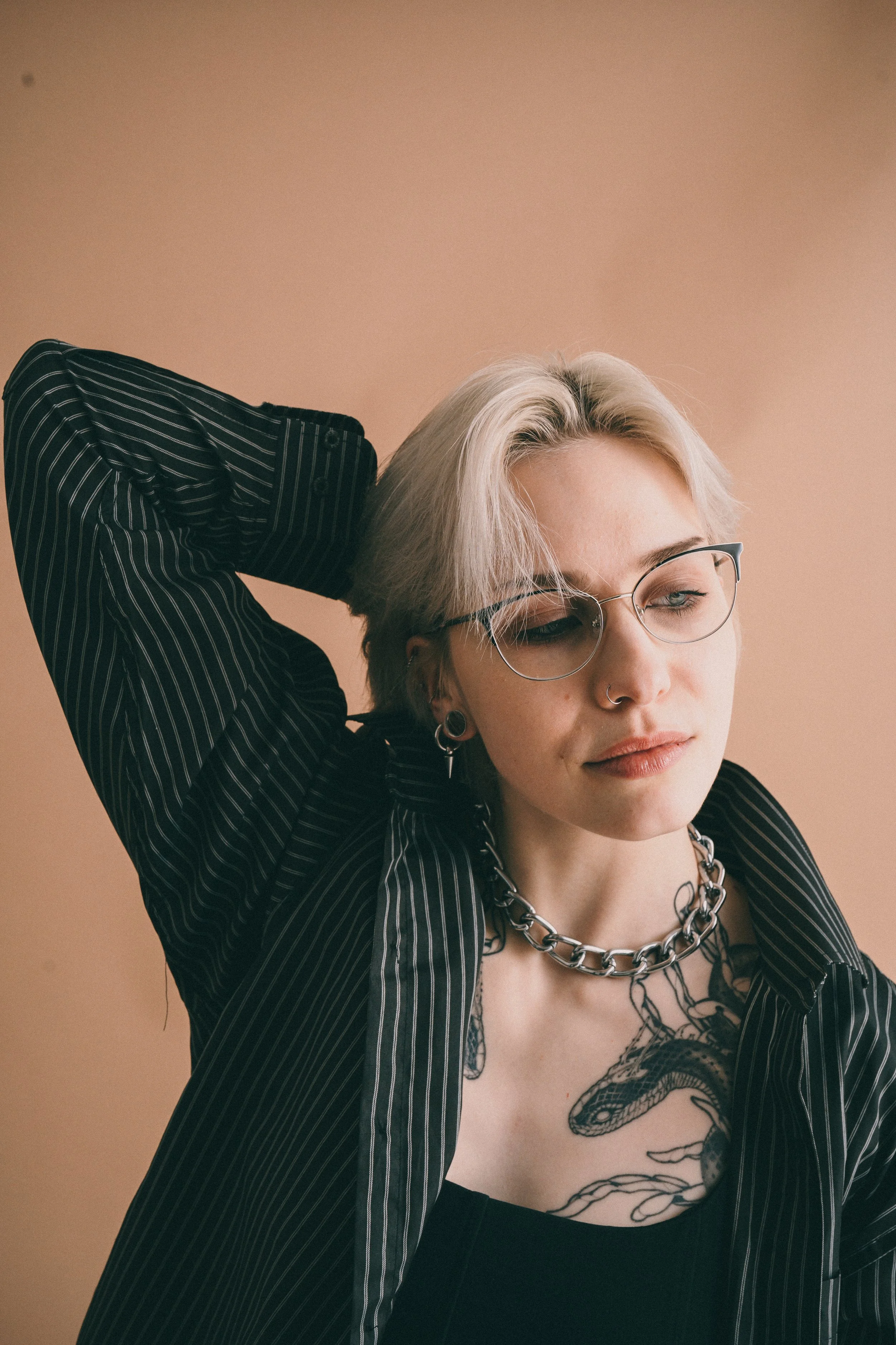 Junge Frau mit Tattoos, schwarze Kleidung, Piercings, kurze blonde Haare, Brille, und Kette vor beige Hintergrund.