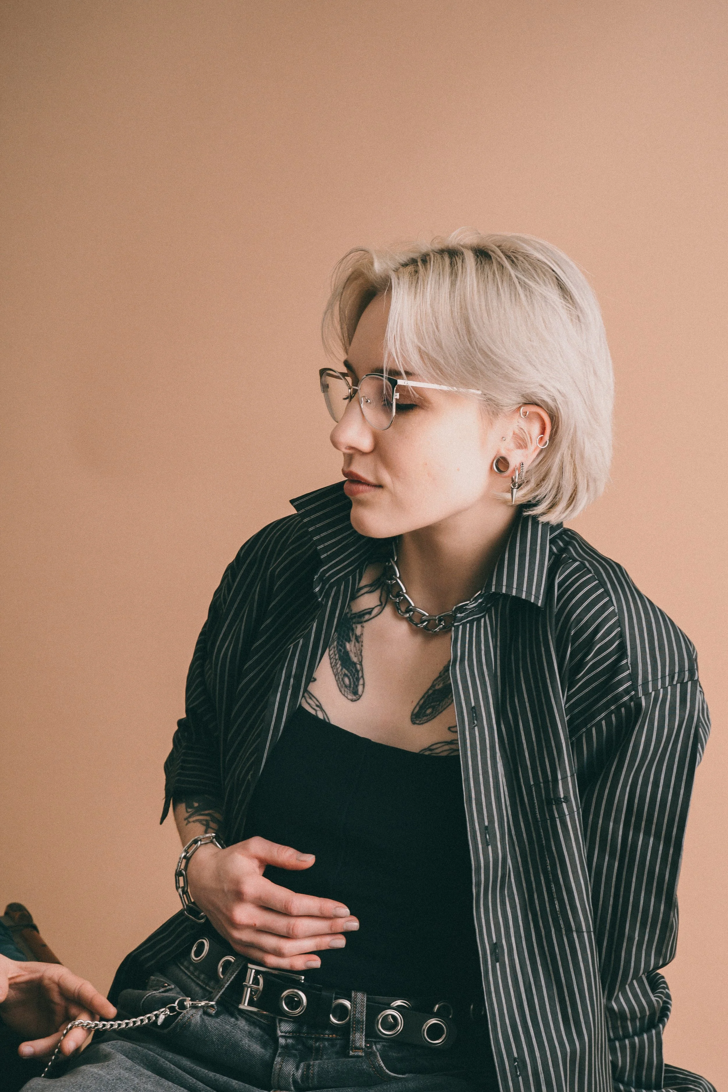Junge Frau mit blondem, kurz geschnittenem Haar trägt schwarze Brille, mehrere Ohrringe und eine Kette, schwarze Lederjacke mit weißen Streifen über einem schwarzen Top, sitzt vor beige Hintergrund.