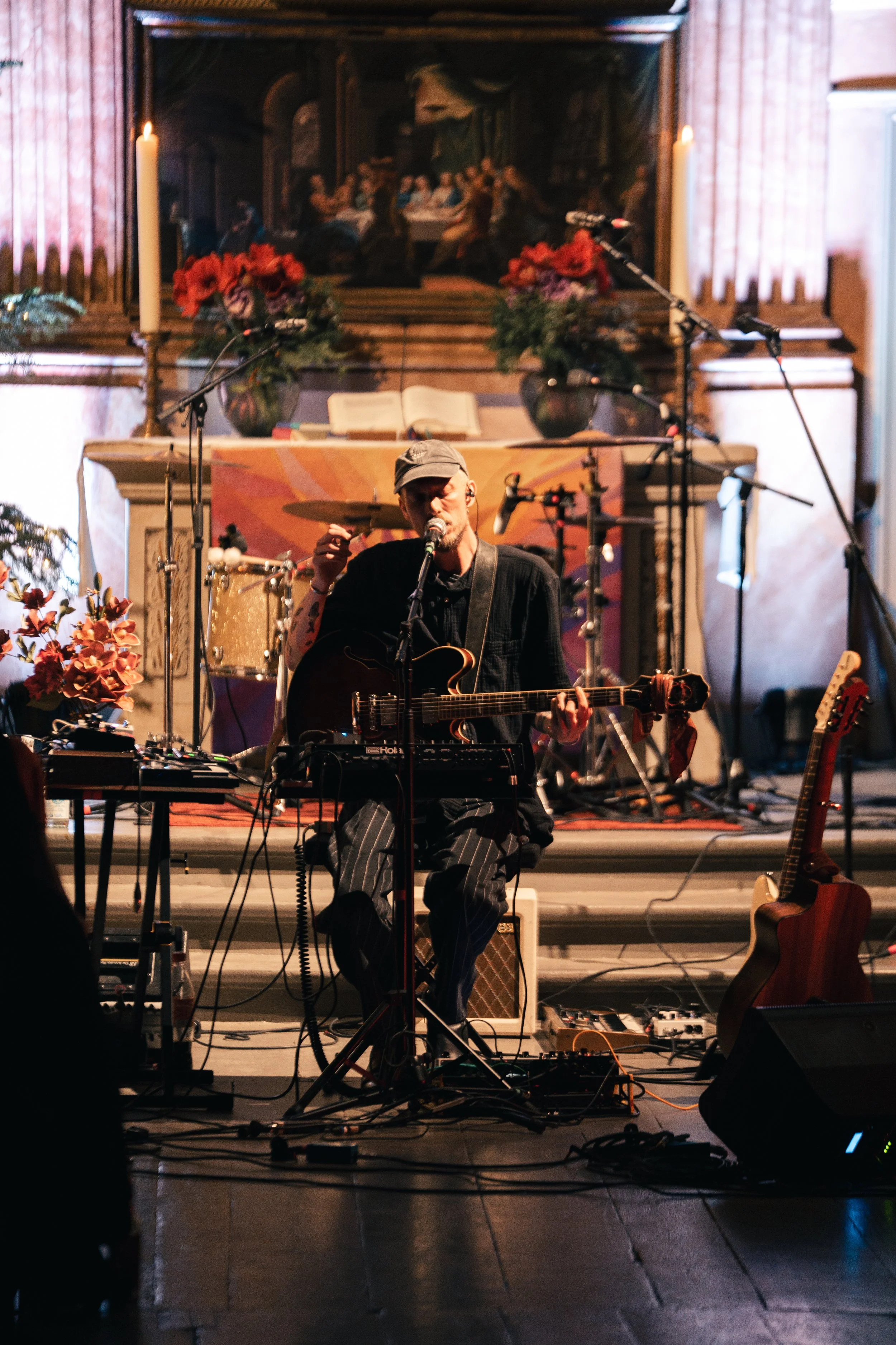 Musiker spielt Gitarre und singt auf einer Bühne mit Mikrofon vor einem Altar mit Kerzen und Blumen; im Hintergrund ein Gemälde einer religiösen Szene.