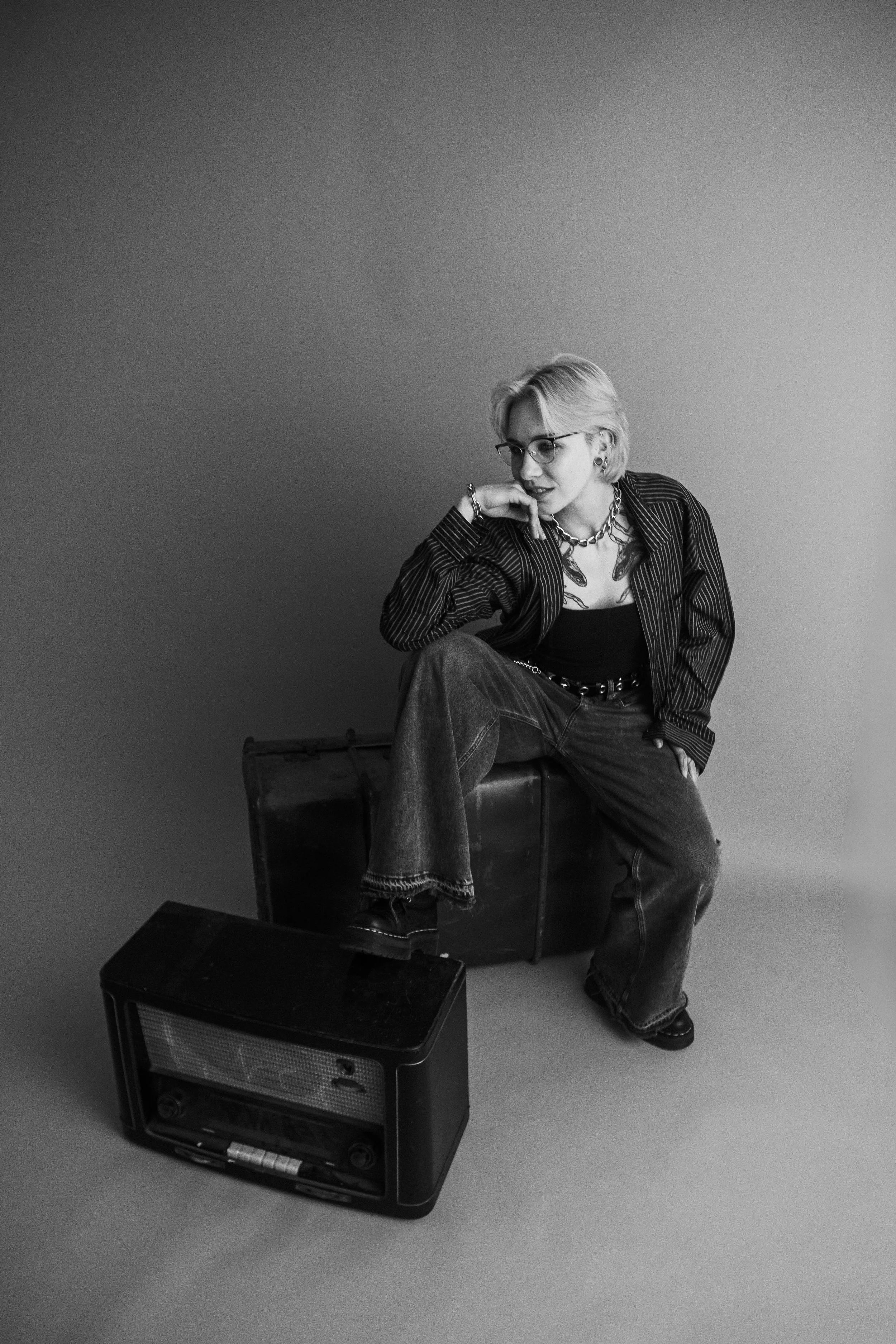 Schwarz-Weiß-Fotografie einer Frau mit kurzen, hellen Haaren, Brille, schwarzer Top, Schmuck und Jeans, die auf einem Koffer sitzt und neben einem alten Radio steht.