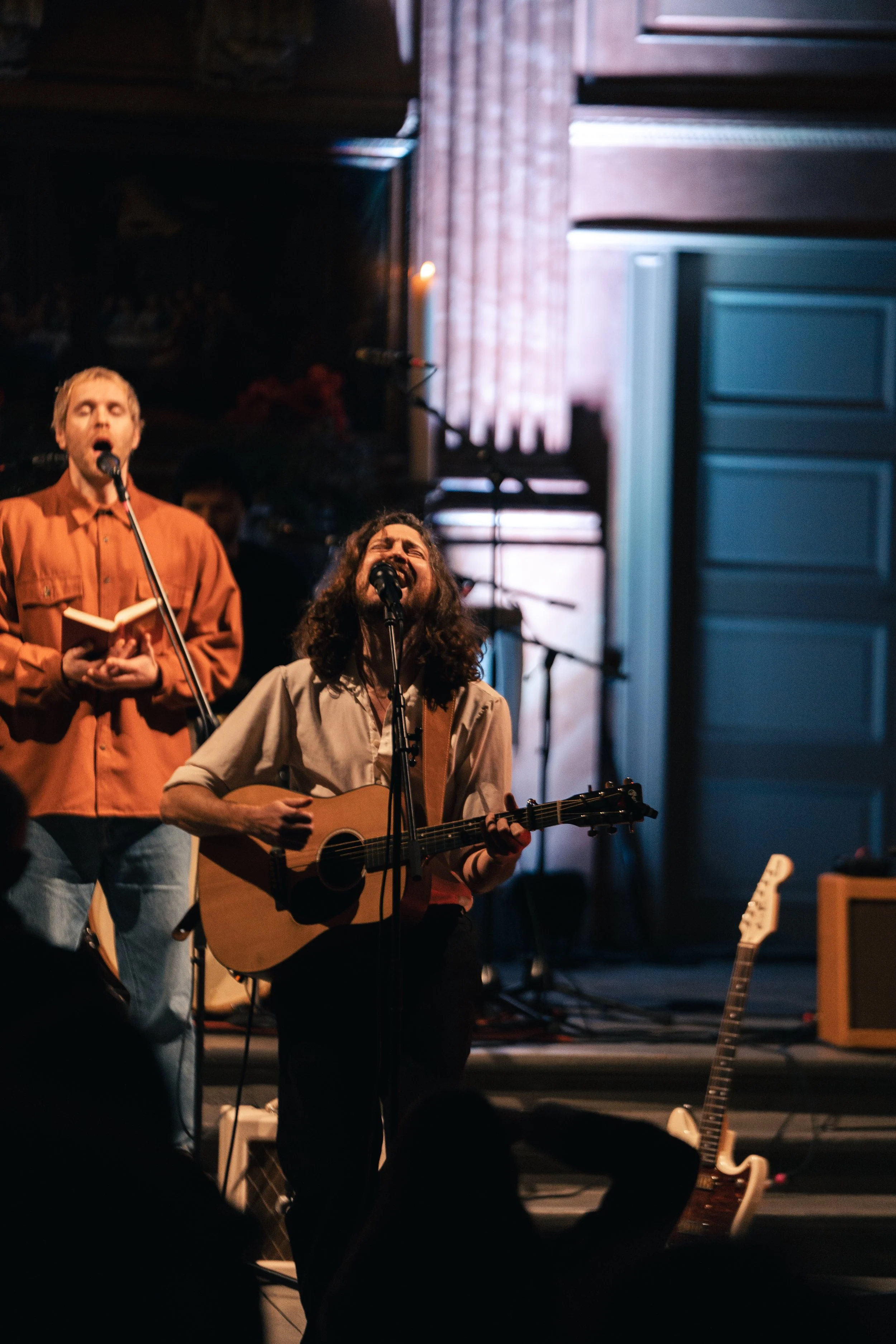 Musiker beim Singen und Spielen einer Gitarre während eines Live-Konzerts, im Hintergrund andere Musiker und Theaterdekoration.