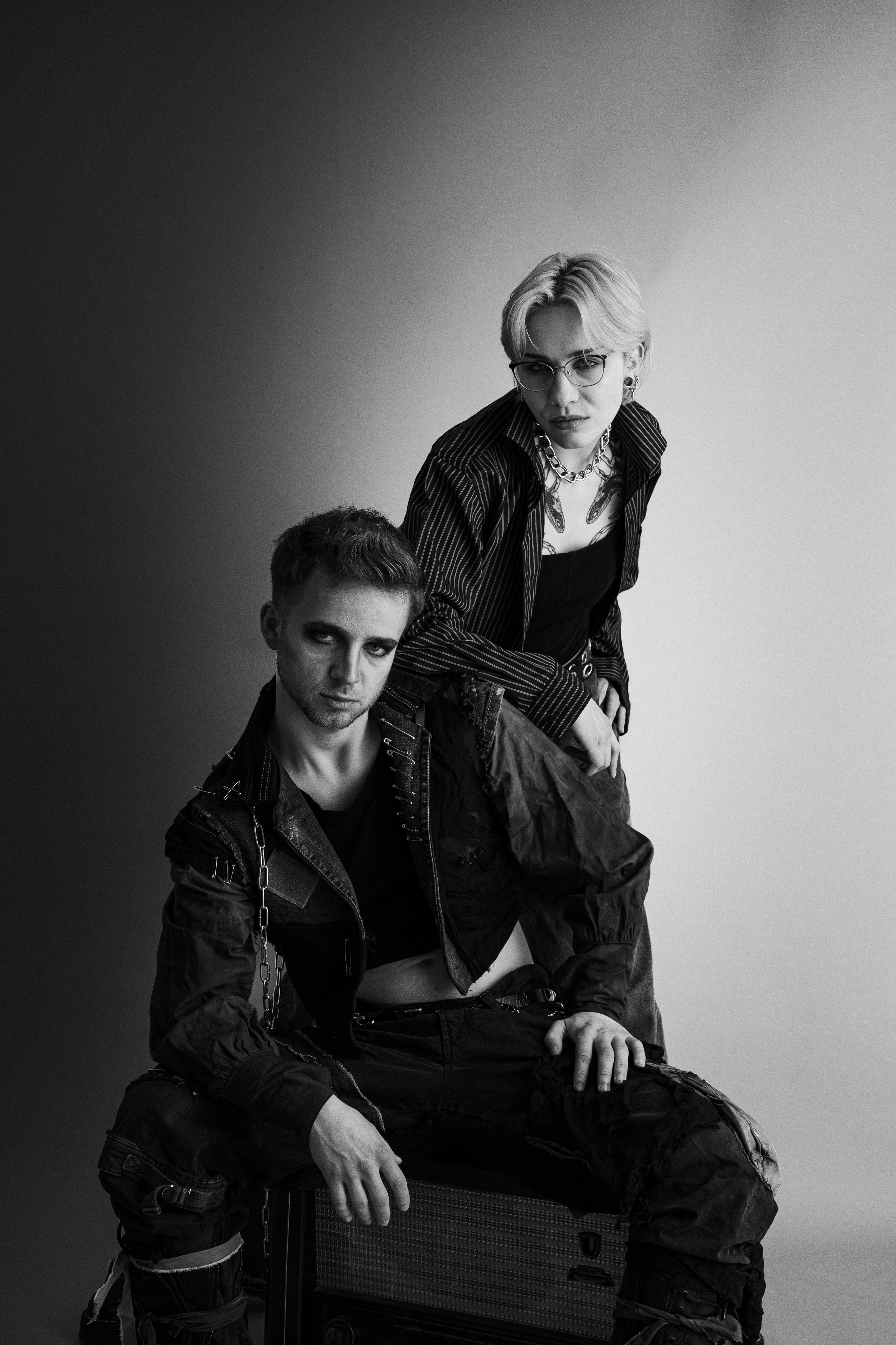 Zwei junge Personen im alternativen Fashionstil, schwarz-weiß Fotografie, einer sitzt auf einem Koffer, der andere steht dahinter, beide schauen ernst in die Kamera.