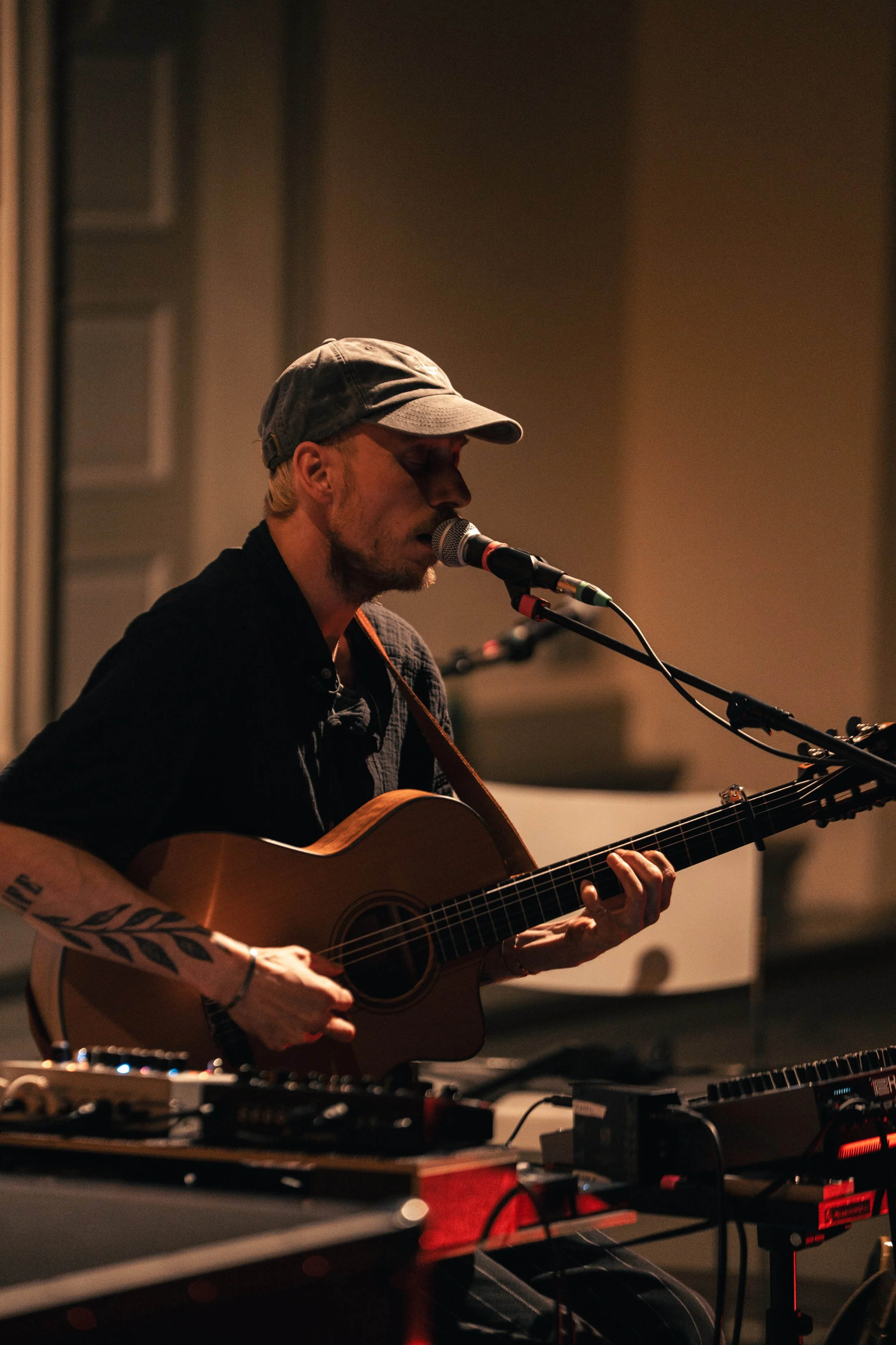 Ein Mann mit Hut spielt Gitarre und singt in einem dunklen Raum, umgeben von Musikelequipment.