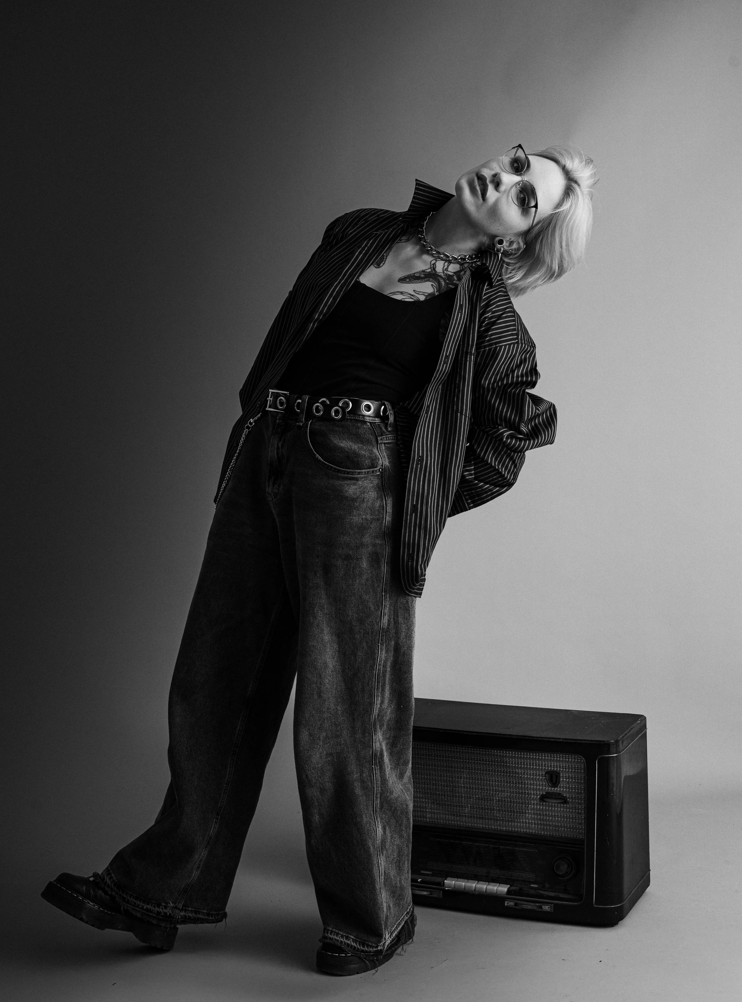 Schwarz-weiße Aufnahme einer jungen Person mit kurzen, hellen Haaren, die eine elegante dunkle Brille trägt, einen Gürtel und eine gestreifte Jacke, neben einem alten Radio, vor einem neutralen Hintergrund.