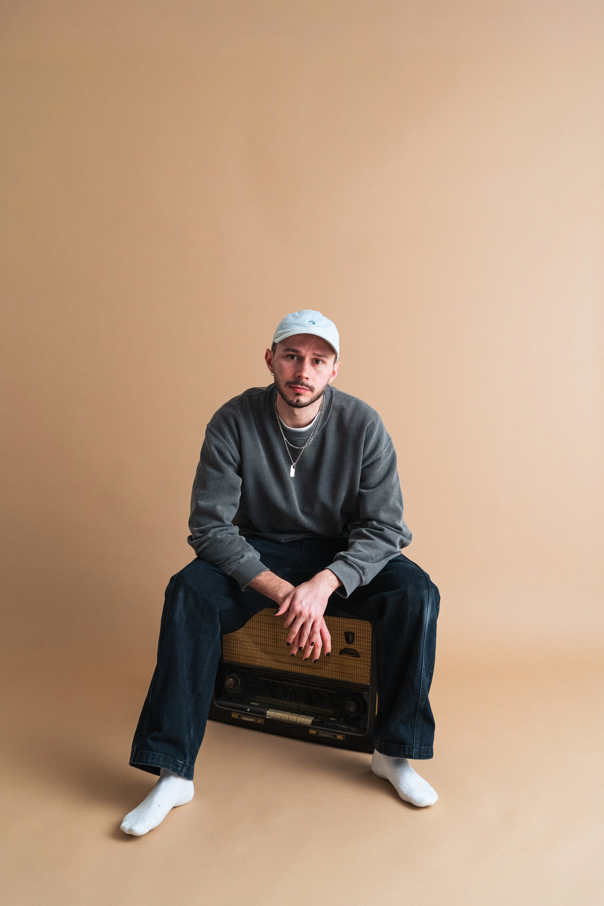 Junge sitzt auf einem vintage Radio vor einem beigen Hintergrund, trägt grauen Pullover, weiße Mütze und dunkle Jeans.