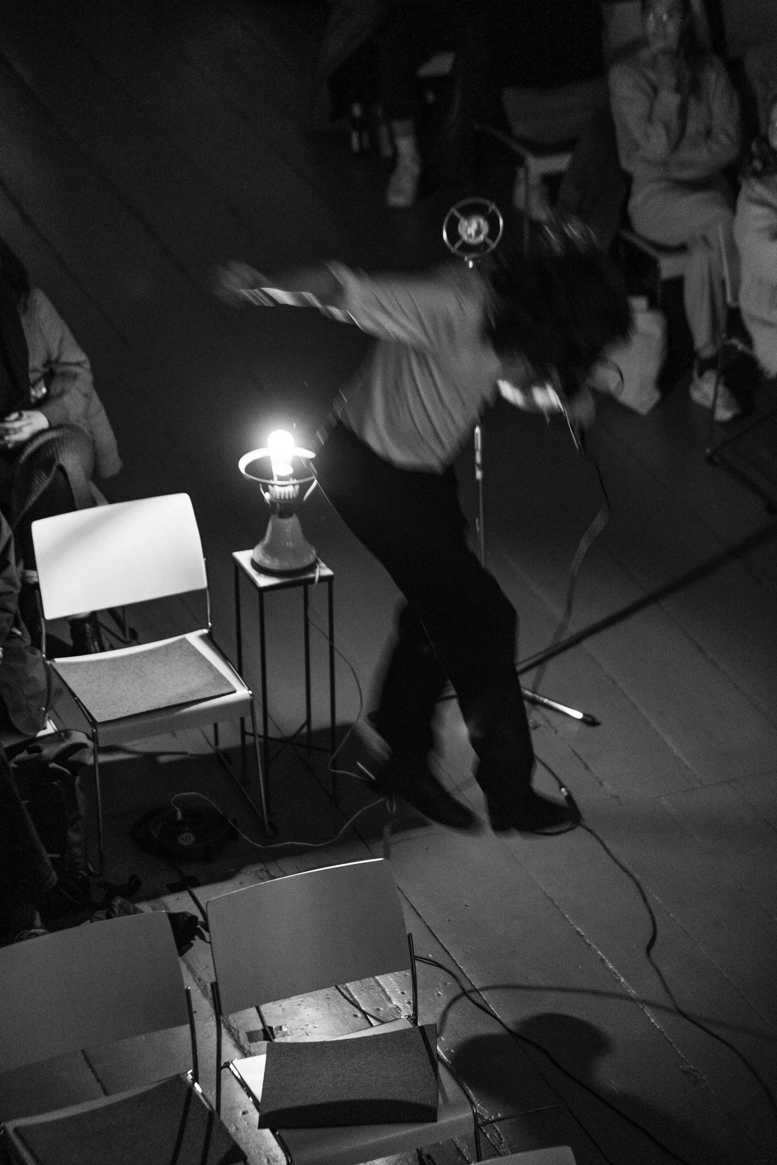 Schwarz-weiß-Foto eines Mannes, der auf einer Bühne tanzt, neben einem kleinen Tisch mit einem Tischlampen-Lampe, in einem Raum mit Sitzgelegenheiten.