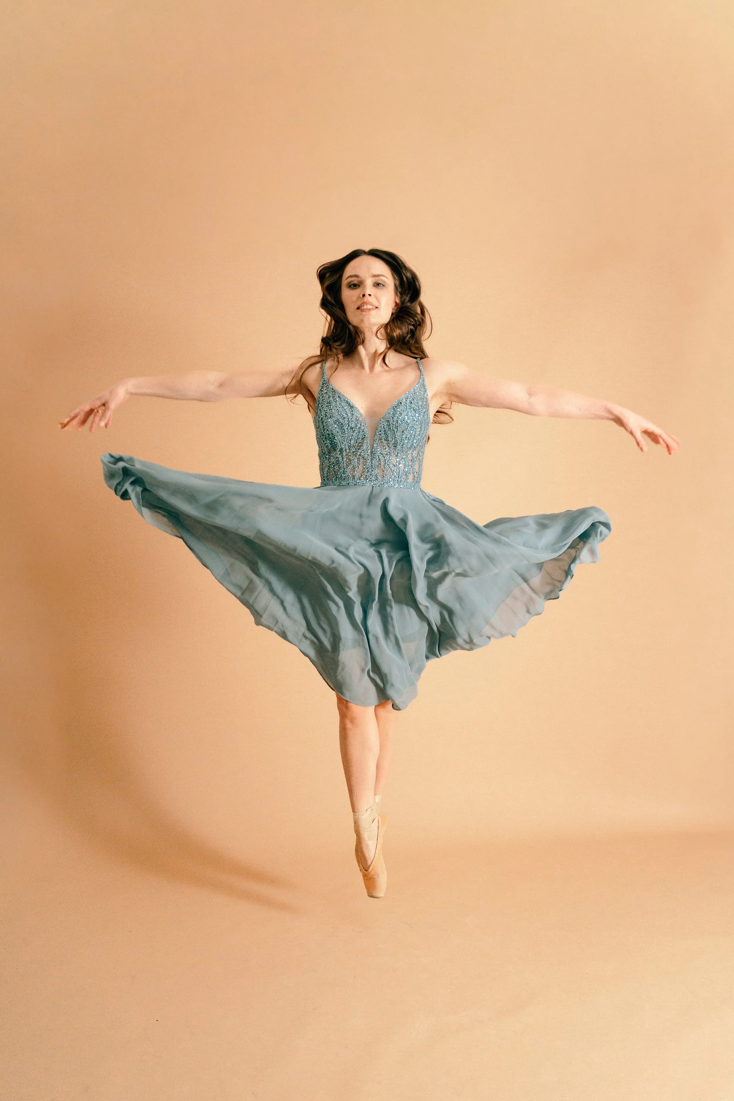Balletttänzerin in einem lightly gekräuselten blauen Tanzkleid, die auf Zehenspitzen tanzt, vor einem hellen, beige Hintergrund.