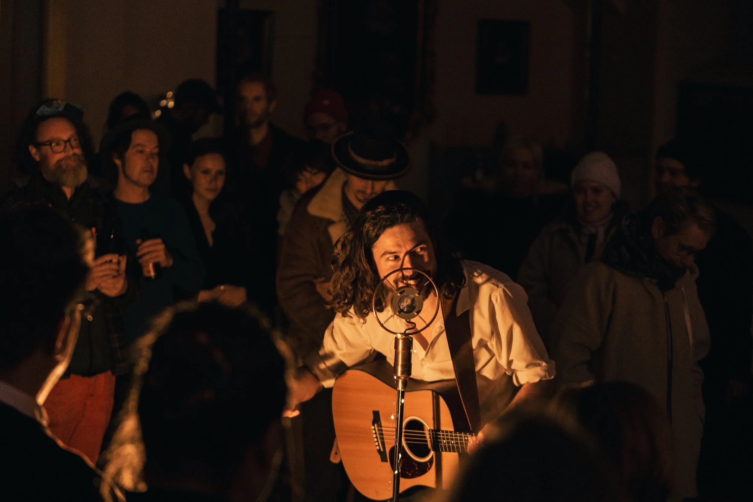Mann spielt Gitarre und singt in einem dunklen Raum, umgeben von einem Publikum, das aufmerksam zuschaut.