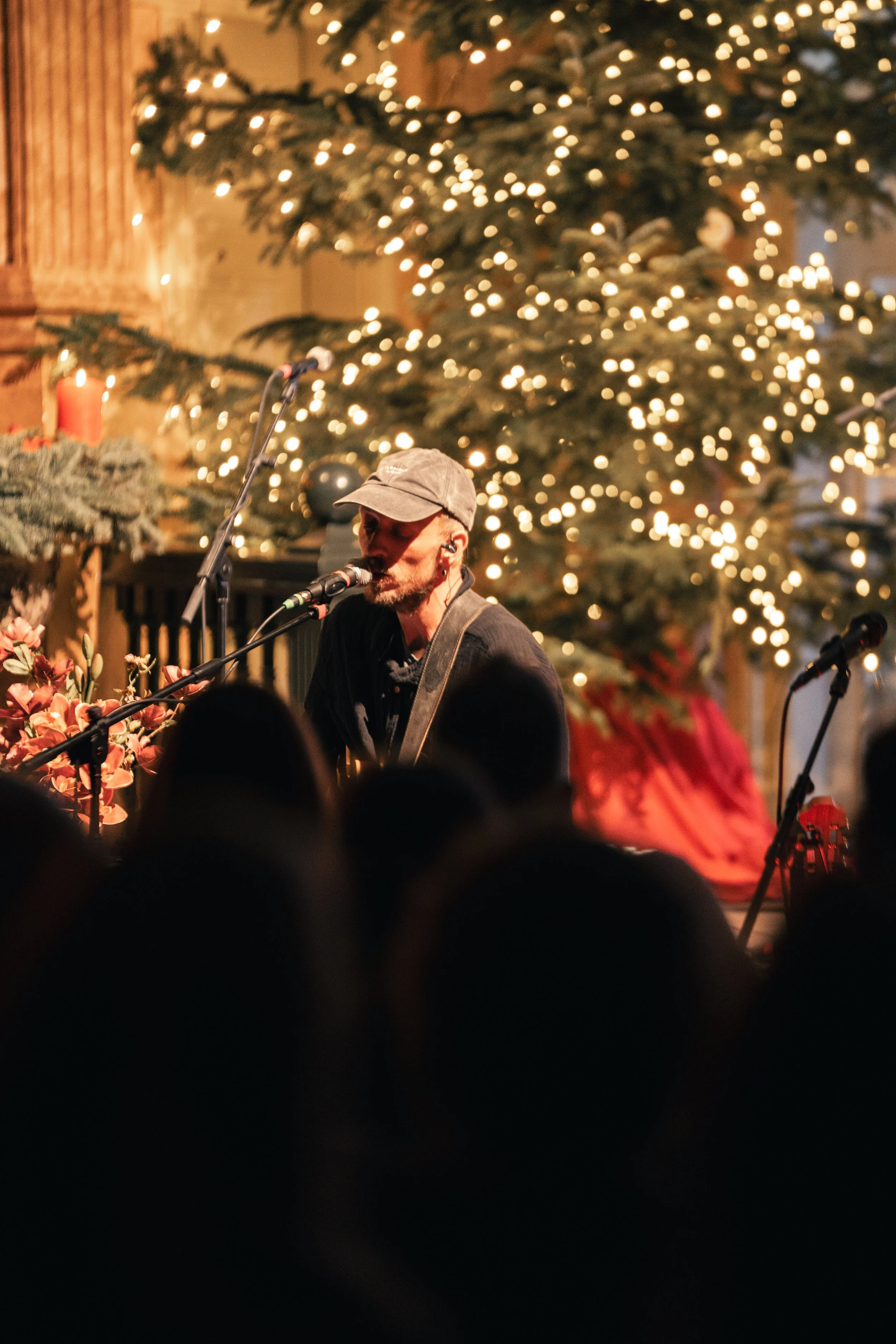 Ein Mann singt mit Gitarre in einer festlichen Weihnachtsumgebung, umgeben von Weihnachtsbaum mit Lichterketten und Dekorationen, vor einem Publikum.