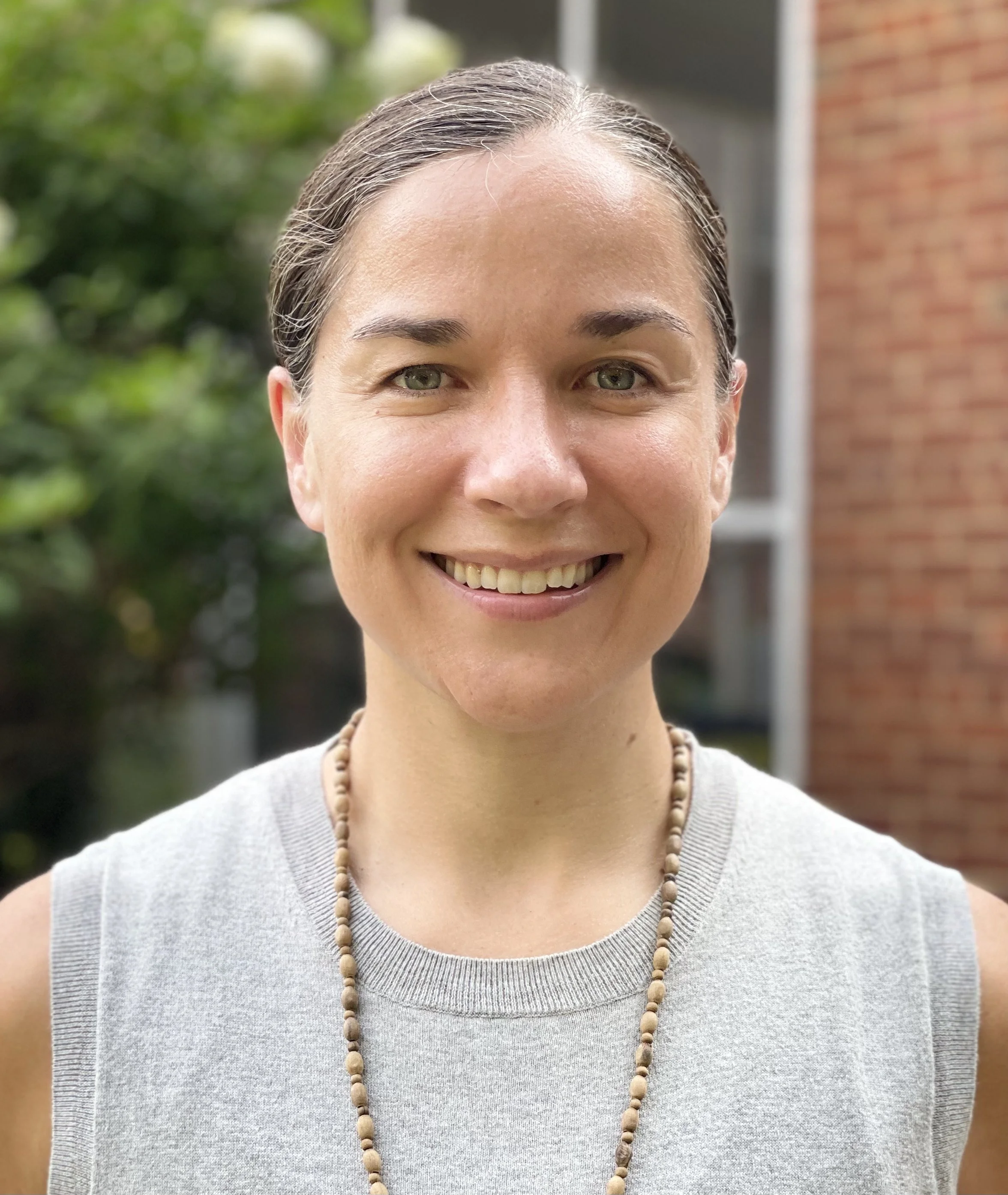 Dr. Meghan Geiss Neuropsychologist smiling, wearing a grey sleeveless top and beaded necklace, outdoors with greenery and a brick building in the background.