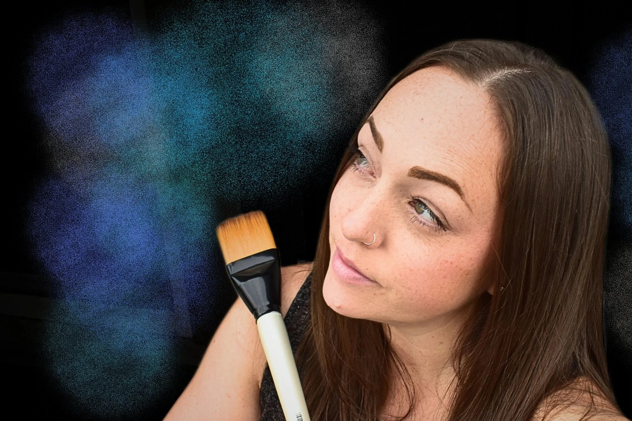 A woman with long brown hair and a nose ring applying paint with a brush against a black background with colorful powder particles.
