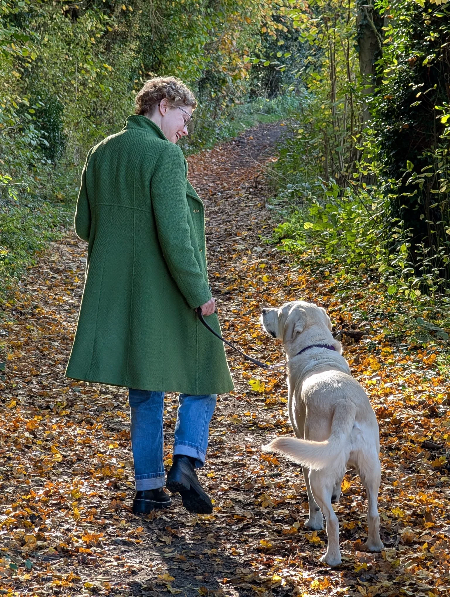 A woman in a green coat and blue jeans walking a yellow Labrador retriever on a dirt trail through a wooded area with fallen leaves.