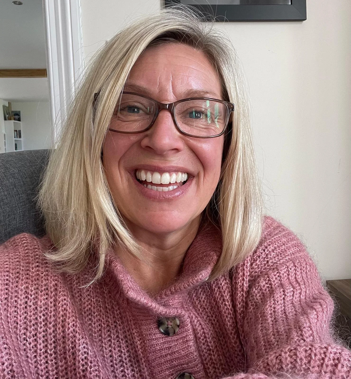 A woman with blonde hair and glasses smiling at the camera, wearing a pink knitted sweater, sitting indoors in front of a plain wall.