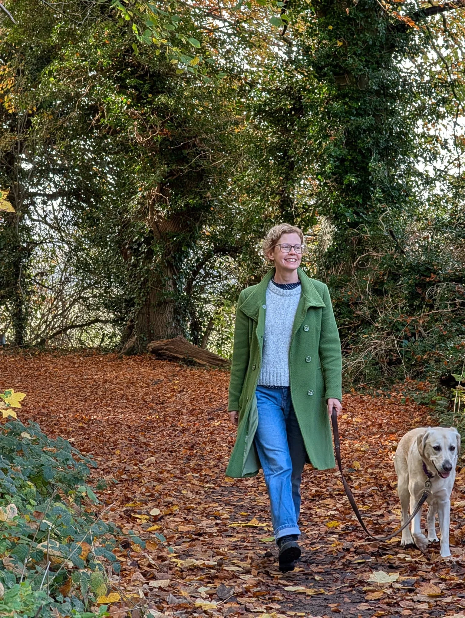 A woman walking her Labrador retriever on a leaf-covered trail in a wooded area during autumn, wearing a green coat, gray sweater, and jeans.