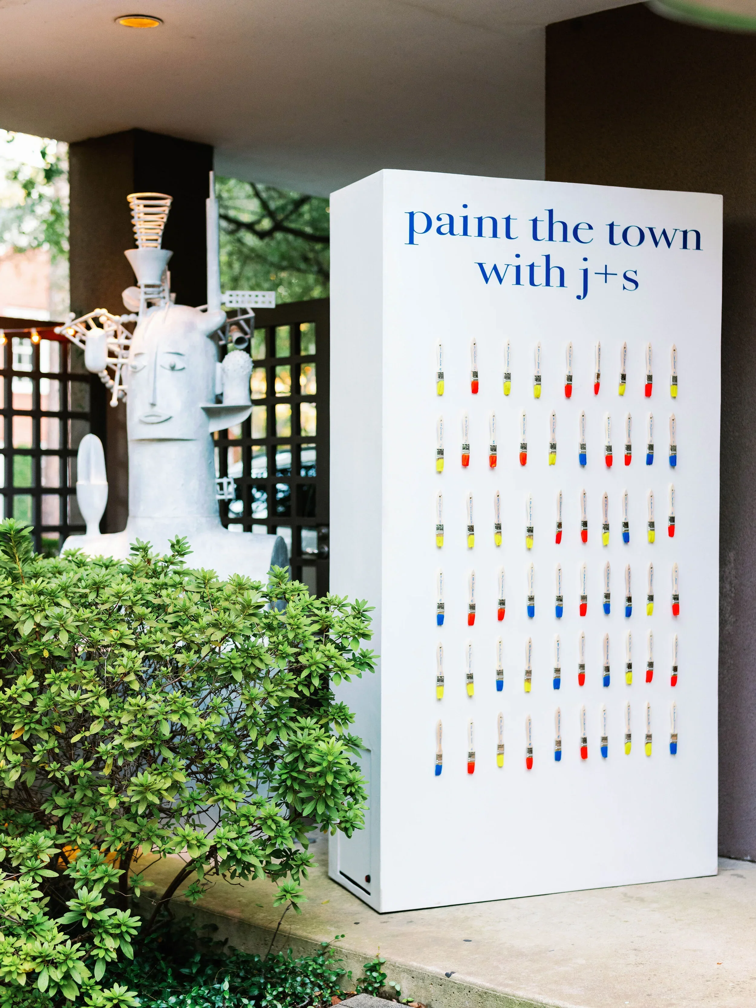 A paint the town with J's display with rows of paint swabs in different colors on a white stand, and a large artistic sculpture in the background.