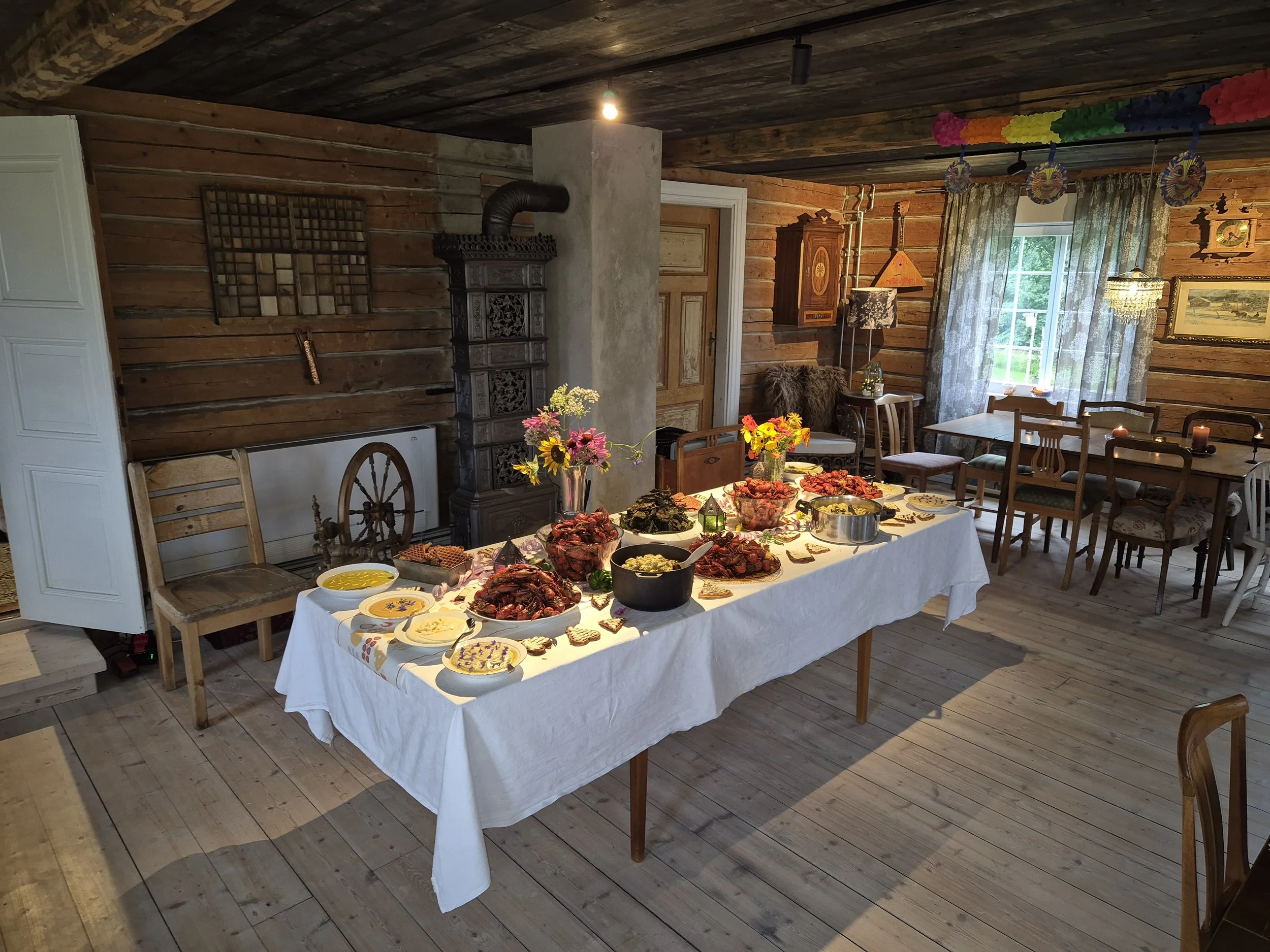 A rustic dining room with wood-paneled walls, set for a meal with a long table covered in a white tablecloth decorated with flowers and numerous dishes of food. There are additional chairs and tables along the walls, and a window with curtains allowing natural light.