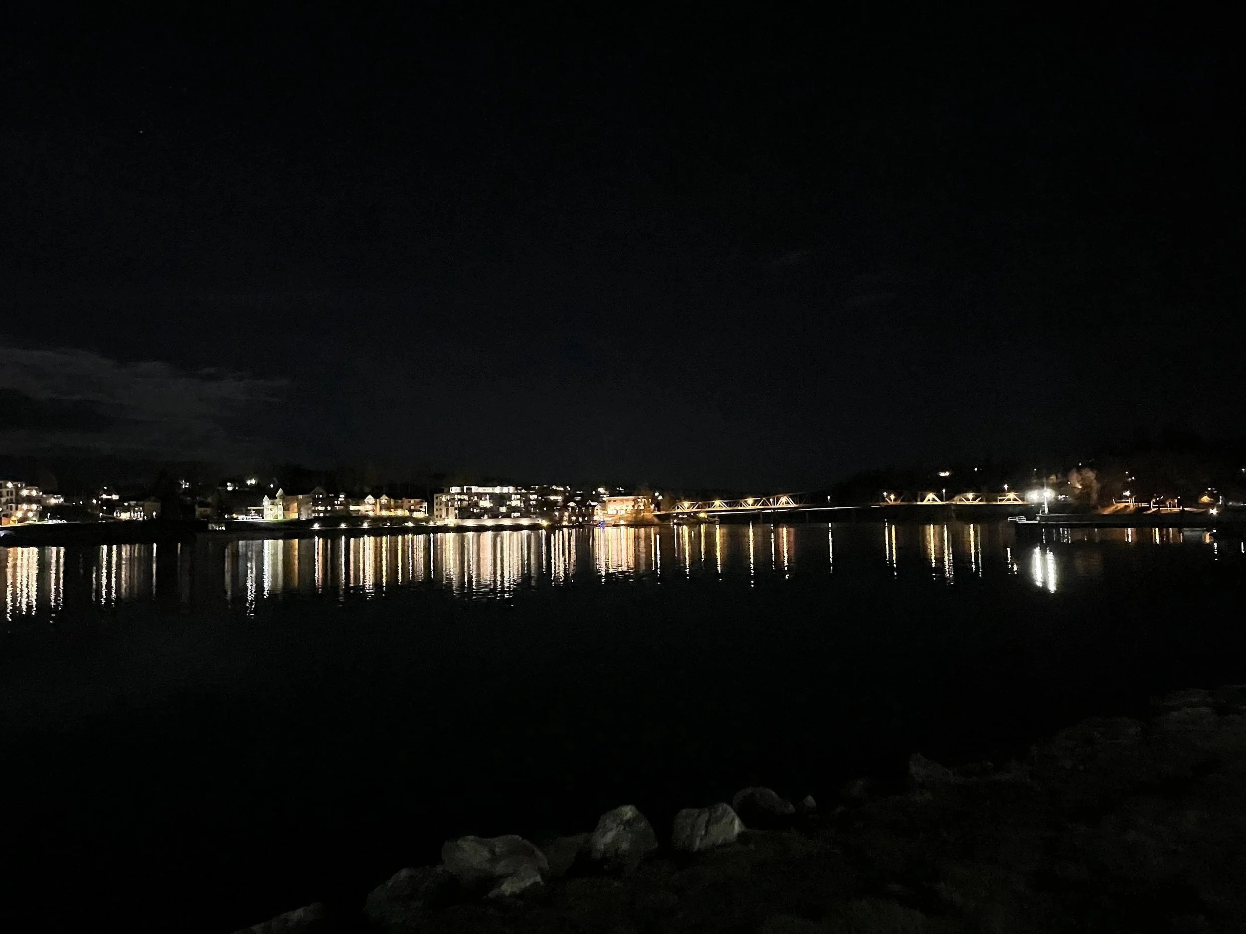 Nighttime view of a lake with reflections of buildings and lights along the shoreline, under a dark sky.
